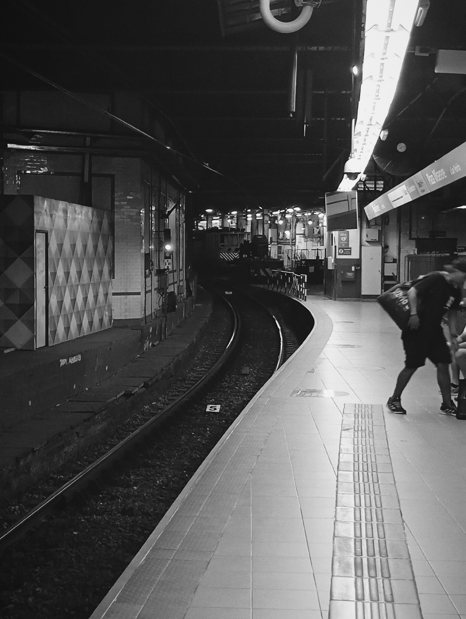 A black and white photograph depicts an underground subway station platform and tracks at Plaza Miserere station, on Line A of the Buenos Aires Subte, Argentina. The perspective is from the platform, looking forward along a curved section of track that bends left.
On the left side of the tracks, a wall with a prominent diamond-patterned relief and a door is visible, alongside various utility conduit and a glowing orange signal light positioned over the tracks. A white marker with the numeral "5" rests on the trackbed. Further in the distance, the front section of a train, identifiable by diagonal stripes, is dimly visible on the tracks.
The platform on the right side of the image features light-colored floor tiles, incorporating a section of tactile paving running parallel to the tracks. Overhead fluorescent tube light fixtures illuminate this area. A station sign, prominently displaying "Plaza Miserere" and other Line A station names such as "Perú," "Piedras," "Lima," and "Congreso," hangs above the platform.
In the foreground on the right, a person is captured in motion blur, walking away from the camera along the platform edge. This individual wears a dark t-shirt, dark shorts, and dark sneakers, carrying a large, dark duffel bag over their right shoulder. The lower legs of a second individual are partially visible adjacent to the walking person, suggesting an additional presence on the platform. The overall environment is functional and typical of a subterranean transit system.