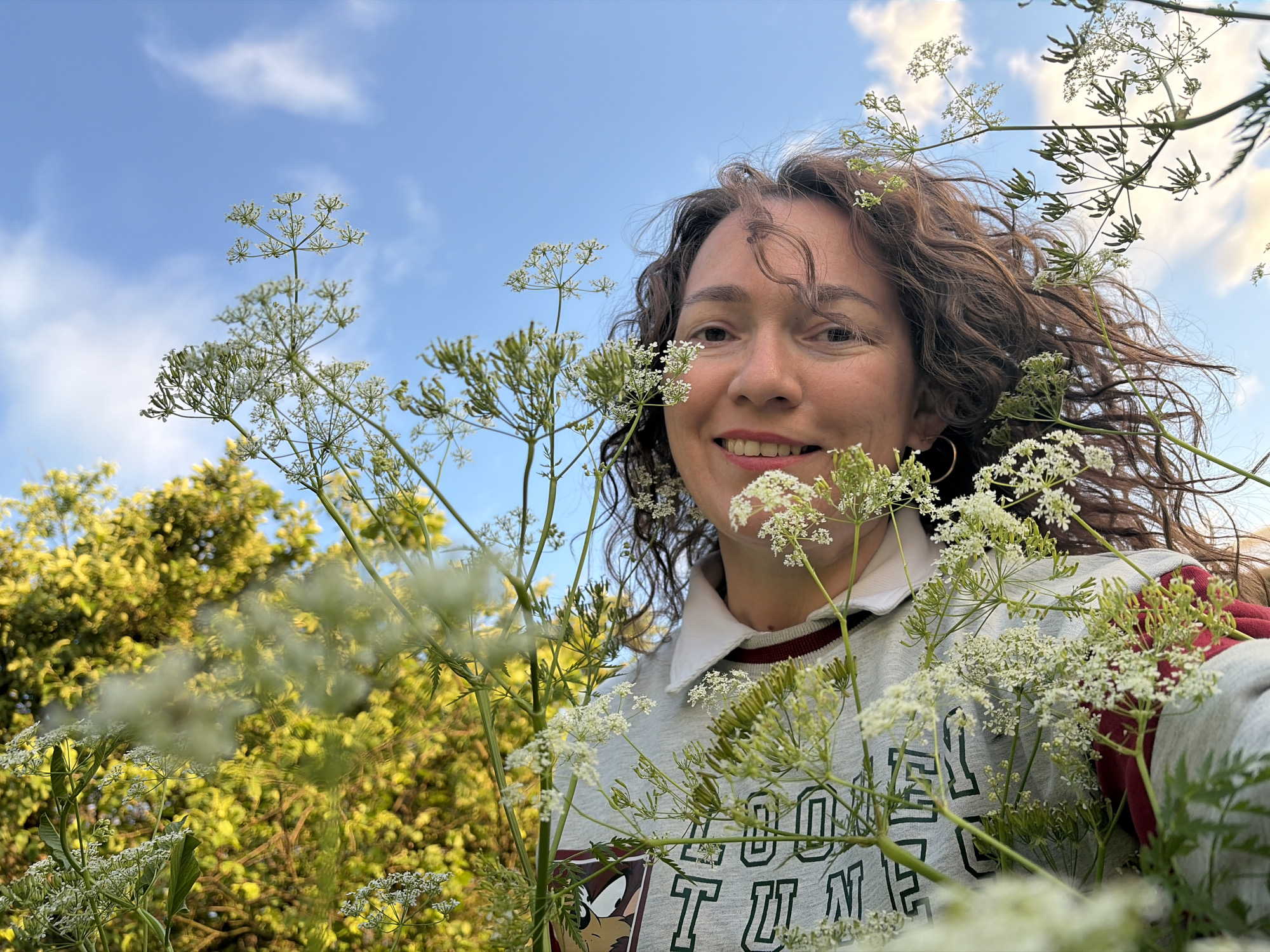 A low-angle, close-up shot shows a woman with curly brown hair smiling at the camera, partially obscured by white Queen Anne's Lace flowers. She is wearing a grey sweatshirt with a white collar and red trim, and a maroon layer underneath. In the background, lush green foliage and a blue sky with wispy clouds are visible. The scene appears to be outdoors, possibly in a field or park, in Olkhovka, Russia.