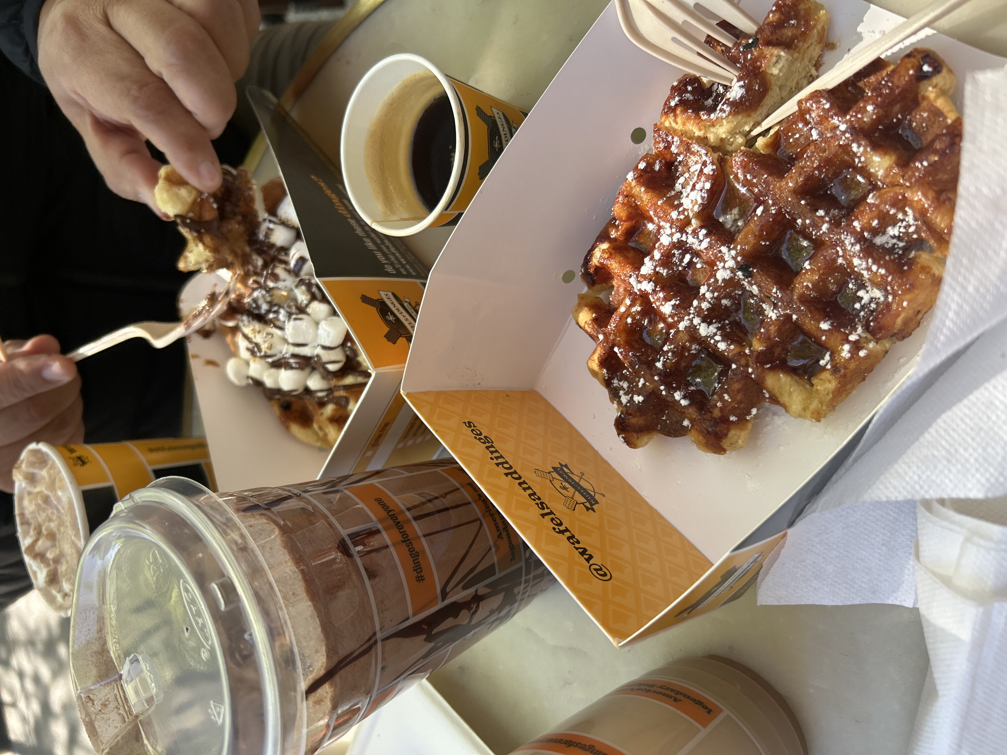 The image captures a bright, high-angle view of a casual meal featuring waffles and drinks from "Wafels & Dinges" in New York, United States of America. In the foreground, a person's hands are visible, one holding a fork to lift a piece of a waffle topped with chocolate sauce and mini marshmallows.
The scene displays two open cardboard boxes, each containing a large Liege-style waffle. One waffle is generously covered in a glossy glaze, powdered sugar, and small green candied pieces, with a plastic fork inserted into it. The other, partially eaten, is adorned with chocolate drizzle and white mini marshmallows.
Several beverages accompany the waffles: a small paper cup containing dark coffee, and a larger clear plastic cup with a lid, showing internal streaks of chocolate syrup, likely a hot chocolate or iced coffee. Another smaller, opaque cup is partially visible. A white paper napkin is also seen tucked into one of the waffle boxes.
The packaging is distinctly branded with "WAFELS & DINGES," a well-known New York establishment. Visible text includes the Instagram handle "@wafelsanddinges," the website "wafelsanddinges.com," the brand name "WAFELS & DINGES" with its logo (a waffle iron and chef), and phrases such as "Do you like these dinges?", "Legendary Belgian Wafels," and "#dingesforeveryone."
The strong natural sunlight and crisp shadows suggest it is midday or early afternoon on a clear, sunny day. The disposable packaging and outdoor setting indicate a casual dining experience, likely at a food truck, stand, or outdoor cafe.
