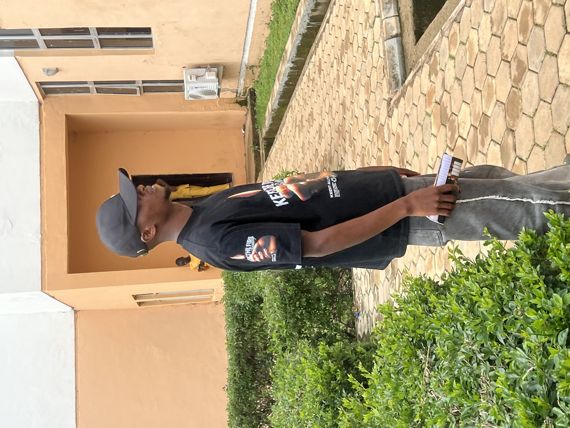 A young man wearing a black t-shirt and a grey baseball cap stands outdoors. The t-shirt has a graphic print on the sleeve featuring a hand and text. He is holding a smartphone in his right hand. The background shows a building with an orange facade, a tiled pathway, and lush green bushes. An air conditioning unit is mounted on the building wall. The scene appears to be in a sunny, daytime environment. The man's expression and posture suggest a casual, relaxed demeanor. The overall setting is an urban or semi-urban area in Wumat, Nigeria.