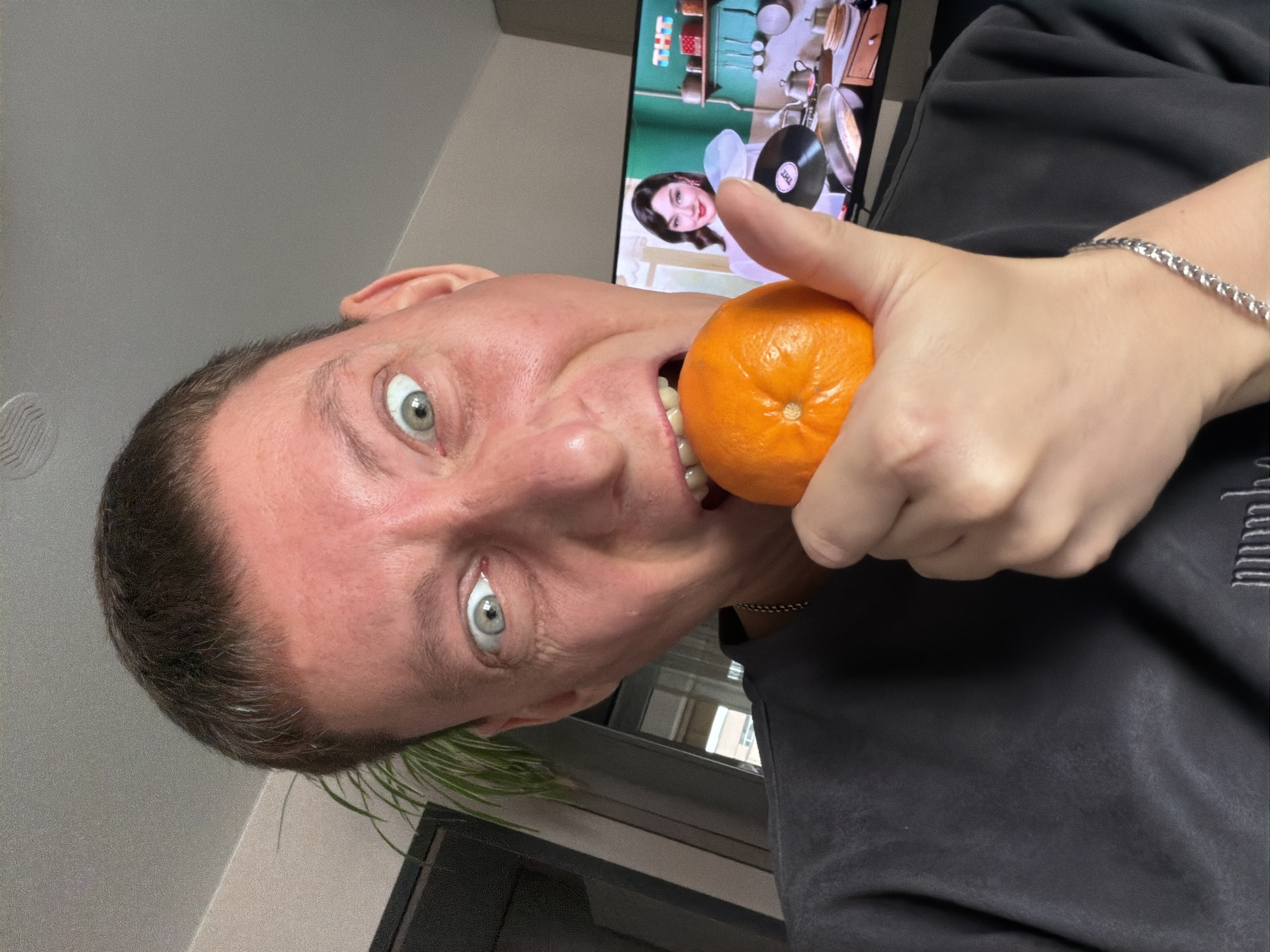A close-up, selfie-style, over-the-shoulder shot shows a person with short, dark hair and wide, pale green eyes holding a tangerine to their mouth. The person is wearing a dark gray t-shirt and a silver chain necklace. Their right hand is holding the tangerine, with their thumb on top and fingers underneath, positioned to bring it to their teeth. The background shows a television screen displaying a woman in a kitchen setting with retro decor, and to the left of the screen is a tall, leafy green plant. The lighting suggests an indoor environment. The scene does not provide any specific details or context to determine the location in the city of Moscow, Russia.