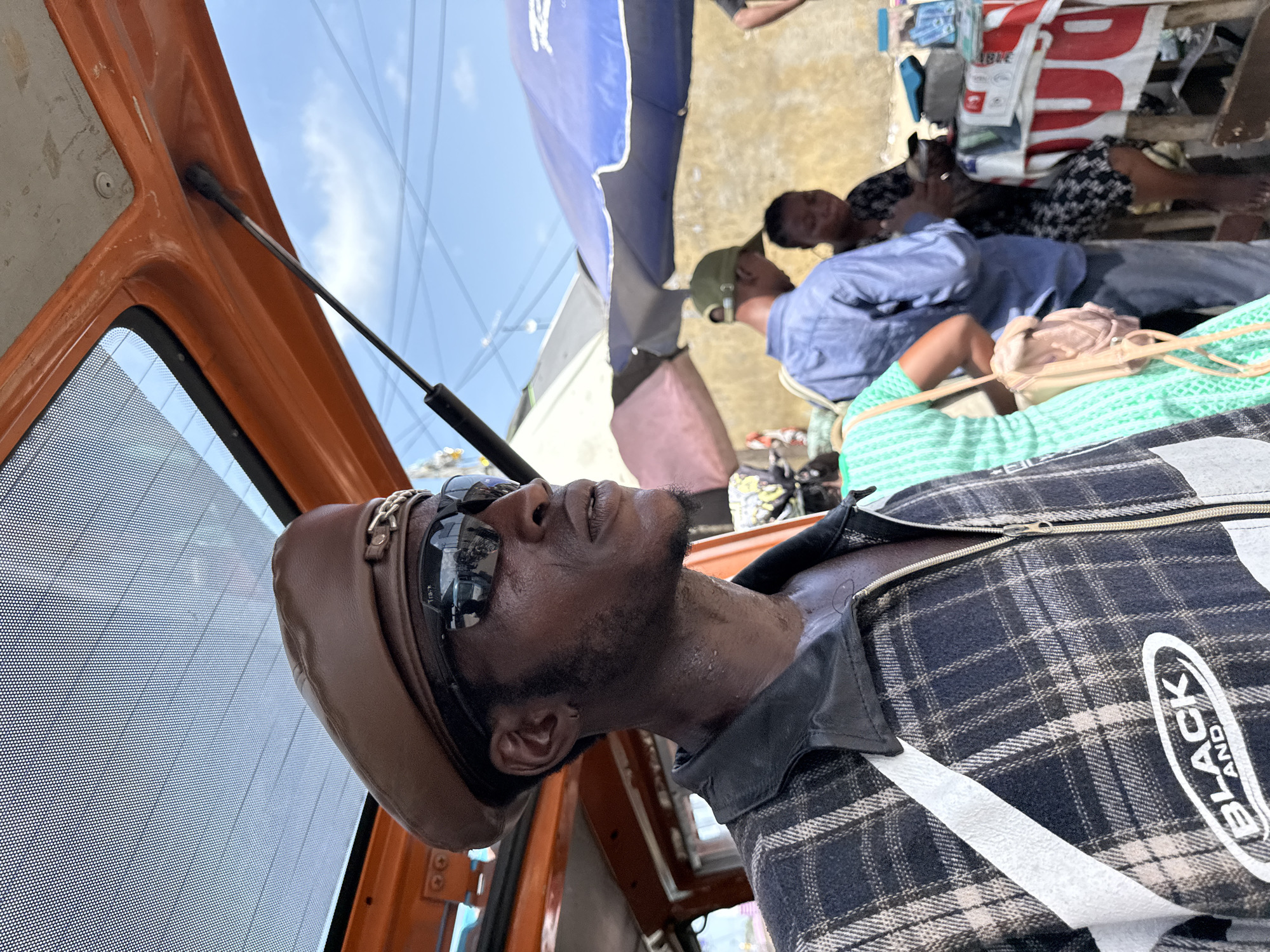 In a candid, high-angle perspective shot taken in Lekki, Nigeria, a person wearing a brown beret and sunglasses is seen inside what appears to be an orange taxi or vehicle. The individual is wearing a plaid jacket with white stripes and the word "BLACK" is visible on the right side of their jacket. In the background, under a blue tarp, there are several individuals, possibly interacting or resting, including one wearing a green baseball cap. The setting seems to be a busy, outdoor environment with an awning visible and a building in the distance.