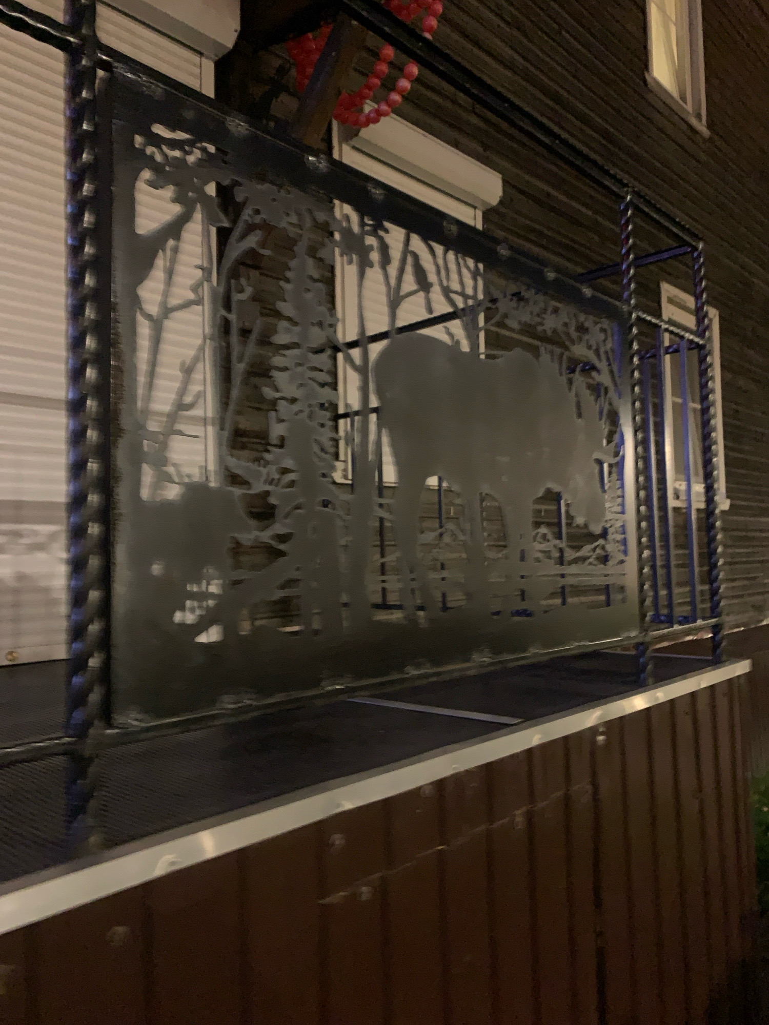 A metal railing with a decorative cutout of a forest scene featuring a moose and a smaller animal, possibly a deer, is visible. The railing is on a balcony or porch of a wooden building. The building's exterior is dark brown wood siding, and there are white shutters on the windows. In the background, there are red beaded decorations hanging. The scene is at night, as indicated by the lighting conditions. The image is taken in Arkhangelsk, Russia.