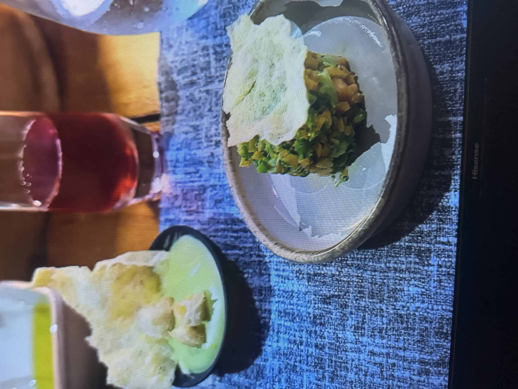 The image shows a close-up of two small dishes of food on a textured blue placemat. To the left, a black bowl contains a pale green dip topped with small, irregular pieces of light-colored food, possibly crackers or croutons. A large, pale yellow, irregularly shaped cracker is propped up against the dip. To the right, a shallow, round, light-colored bowl holds a layered dish of finely diced vegetables, appearing to be orange and green. A piece of pale green food, possibly a cracker or vegetable slice, is placed on top of this dish. In the background, a dark red liquid is visible in a tall glass. The overall impression is of an appetizer or small course. The location in Kyiv, Ukraine cannot be determined from the image.
