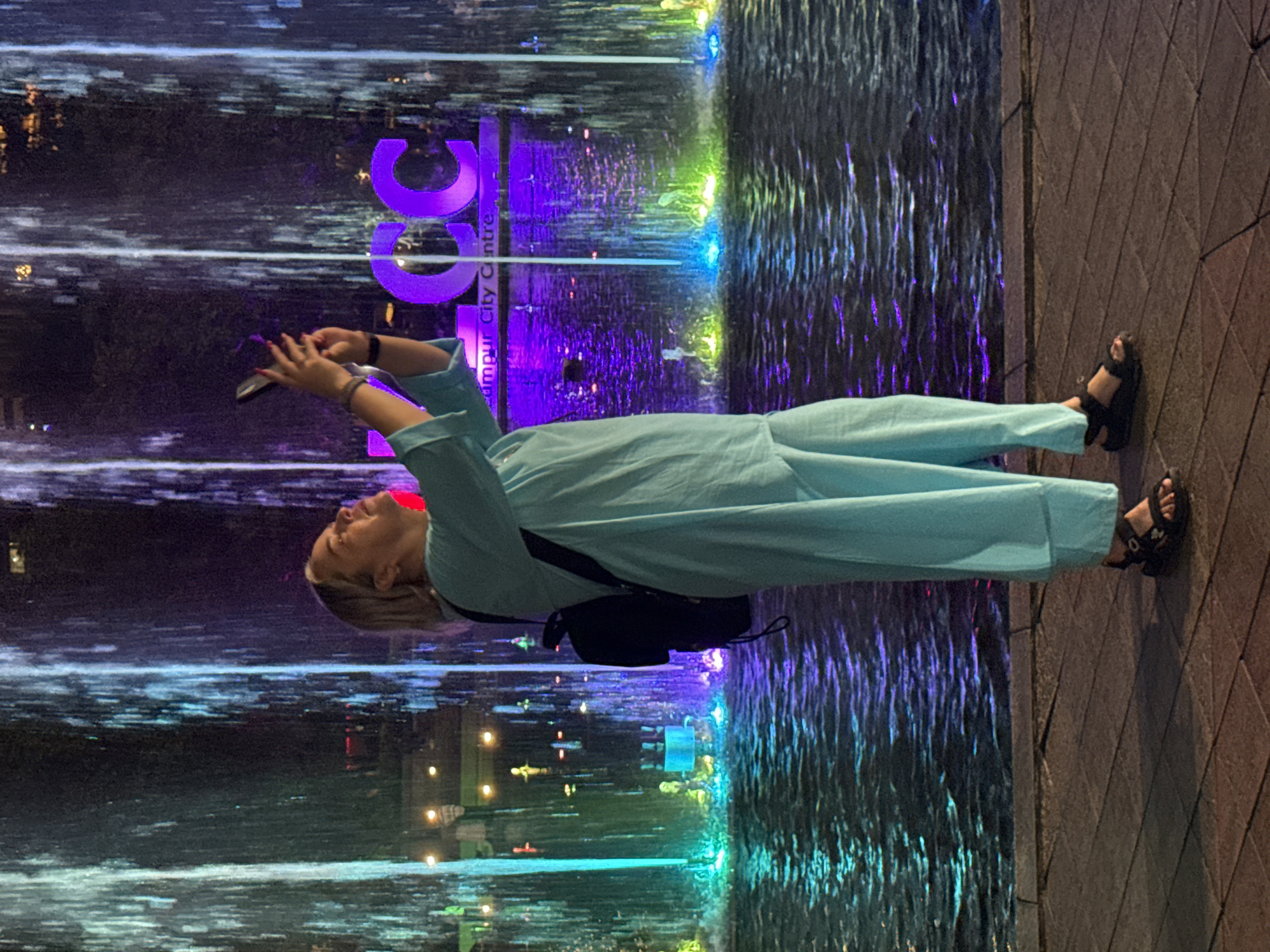The image depicts a woman standing at night in Kuala Lumpur, Malaysia, in front of a brightly illuminated water feature. She is shown from the chest to her feet, wearing a light blue long-sleeved top, matching wide-leg trousers, and black sandals. A black backpack is on her back. The woman has blonde hair and is holding a dark smartphone with both hands, looking upwards as if taking a photo or video of the scene. Her fingernails are painted red.
Behind the woman, a large fountain or waterfall cascades down, intensely lit with vibrant purple, blue, and green lights that create striking vertical patterns and reflections in the water. Through the illuminated water, the large purple letters "KLCC" are prominently visible, with the text "Kuala Lumpur City Centre" written vertically beneath them, identifying the location. To the woman's right, a paved walkway of rectangular tiles is visible. The scene captures the lively atmosphere of a city landmark at night, with the woman engaged in observing and documenting the colorful light and water display.