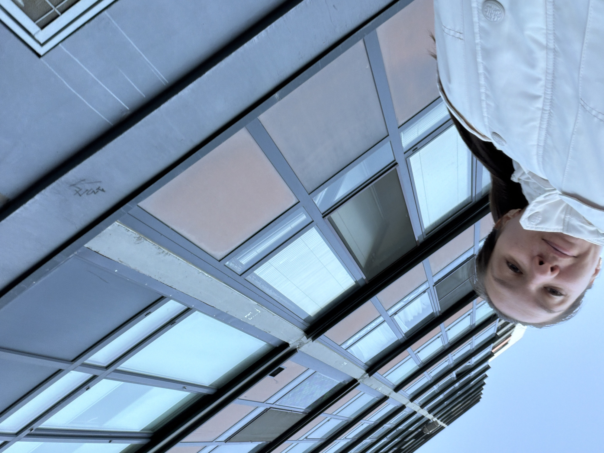 The image depicts a woman in a white jacket standing in front of a tall, modern building in an urban environment. The building's facade consists of a grid of windows and panels. The sky is visible in the background. The location is Unknown, Russia.