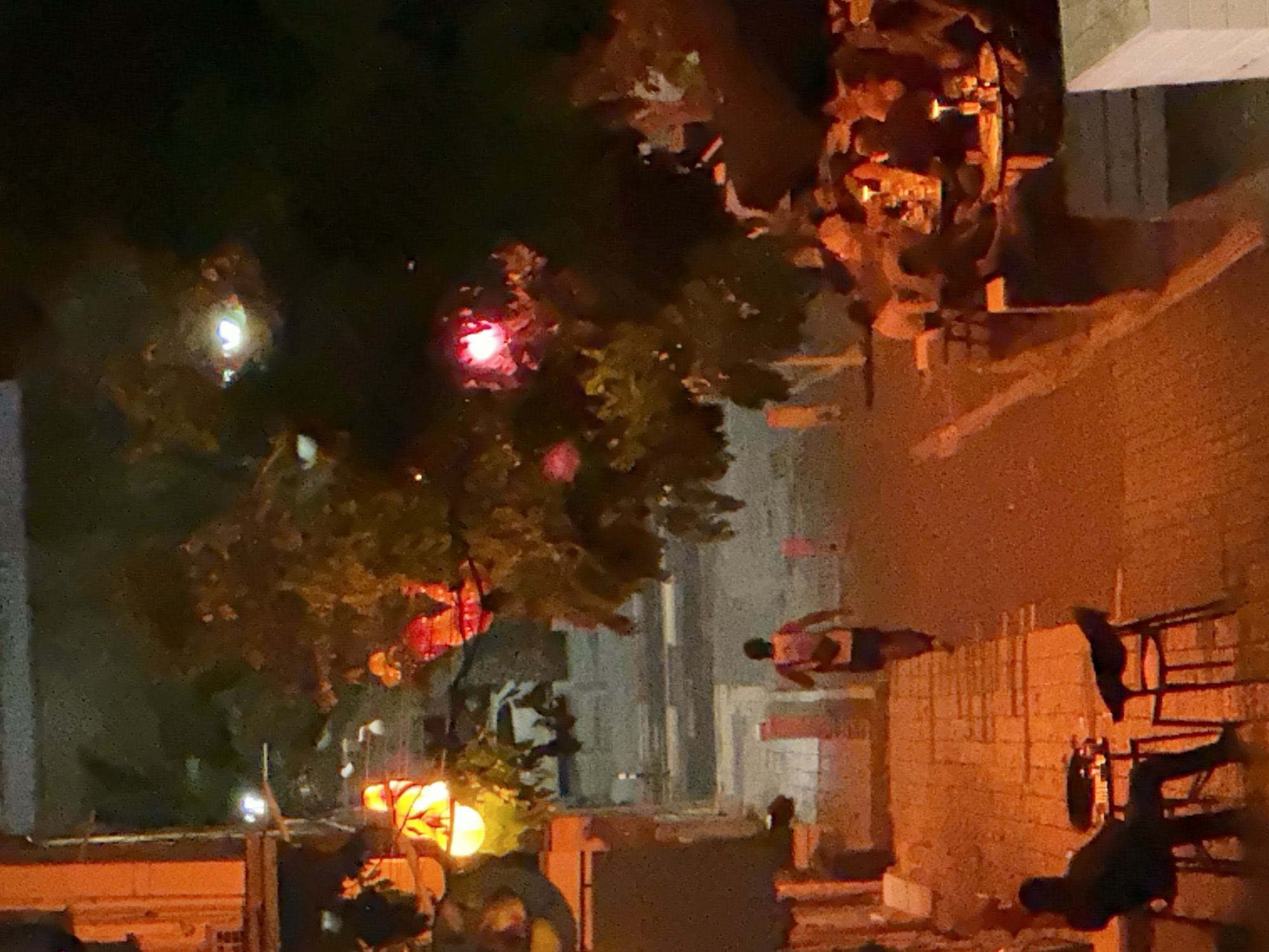 The image depicts an outdoor scene at night, possibly in a city, given the presence of buildings and what appear to be streetlights or decorative lights. The overall lighting is dim, with bright points of light emanating from various sources, including red and orange lanterns or lights, and what seems to be a streetlamp.
Several individuals are visible in the scene. Some are standing and appear to be walking or congregating. One person is wearing a red shirt and dark shorts, and another is wearing a dark top and pants. There are also blurred figures, some of whom appear to be lying down or in a prone position. The presence of multiple people, some of whom are in unusual postures, suggests a gathering or an event is taking place.
In the foreground and mid-ground, there are trees with foliage. Some of the red lights are suspended from or placed amongst the branches of these trees, suggesting a decorative or festive lighting display. To the right of the image, a brick wall is visible, with some structures or scaffolding against it. There are also some tables and chairs that appear to be set up, further indicating a social gathering or event.
The time of day is clearly night, as indicated by the darkness and the artificial lighting. There are no obvious cues about the weather, but the lighting conditions are consistent with a clear night. The location context provided mentions Haifa, Israel.
Notable details include the contrasting colors of the lights, particularly the bright red and orange hues against the dark background. The arrangement of people, especially those who appear to be lying down, could suggest a protest, a performance, or simply people resting. However, without more context, it's difficult to determine the exact nature of the activity. There is no visible text in the image.