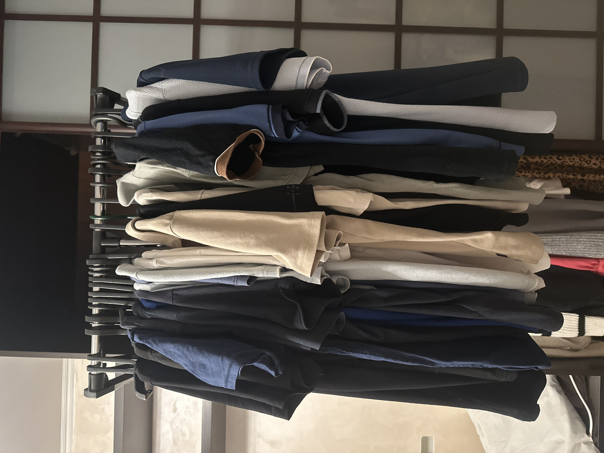 The image shows a rack of men's clothing hanging on hangers. The clothing consists of several t-shirts and polo shirts in various colors including black, navy blue, white, and tan. The rack is positioned in what appears to be a closet or dressing area, with a tiled wall in the background and a wooden screen on the right. There are no people visible in the image. The lighting is dim. The location is not specified in the image or its metadata.