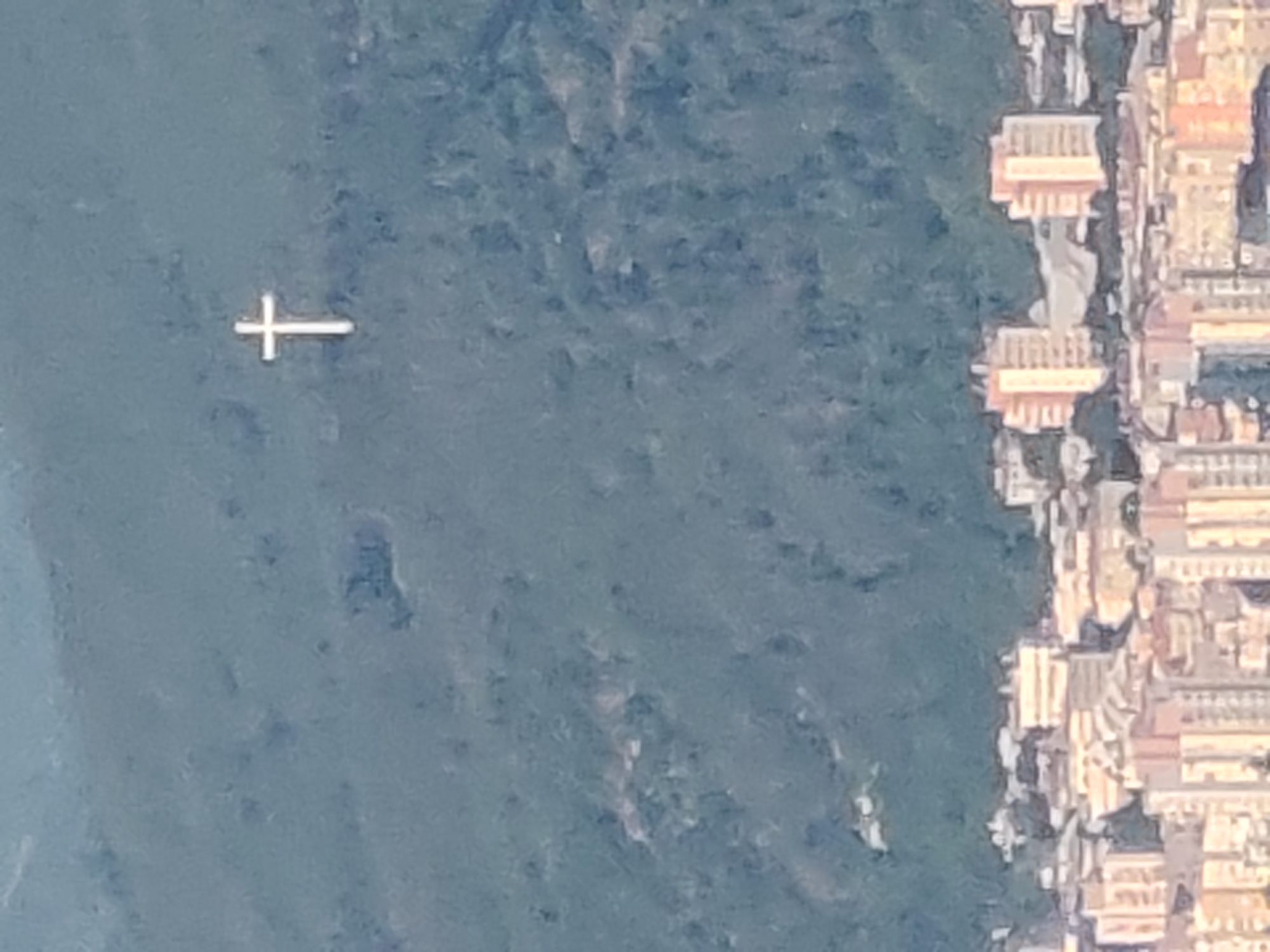 This image presents an aerial or high-angle view of a serene waterfront scene, likely rotated 90 degrees counter-clockwise. On the left, a large, calm body of water occupies a significant portion of the frame, displaying various shades of blue and green, suggesting depth or reflections from surrounding land. A distinct white, cross-shaped object floats on the water towards the upper left, its purpose unspecified but serving as a focal point.
To the right, a densely packed settlement lines the edge of the water. The buildings, mostly light-colored with warm-toned roofs (beige, orange, reddish hues), appear to be residential or small commercial structures, showing multiple stories and typical architectural details such as windows and balconies. They are arranged closely together, following the contour of the shoreline or a path alongside it.
No people are visible, and no specific activities are discernible, leaving the scene tranquil and static. The lighting suggests it is daytime, with clear visibility and subtle shadows indicating sunlight. The weather appears fair with calm water. There is no visible text in the image. Given the context of Selishte, Bulgaria, this scene likely depicts a view over a lake or a wide river within the region, possibly the Iskar Reservoir, with a town or village along its banks.