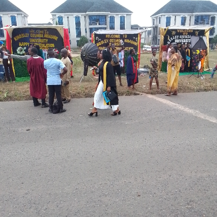 The image displays an outdoor scene at Kingsley Ozumba Mbadiwe University in OGBOKO, Nigeria, during a matriculation ceremony. In the foreground, an asphalt road runs horizontally across the lower half of the frame. Beyond the road, on a grassy verge, three decorated temporary photo booths are set up. Each booth features a banner prominently displaying "WELCOME TO KINGSLEY OZUMBA MBADIWE UNIVERSITY HAPPY MATRICULATION." The booths are adorned with gold, brown, and green fabrics, and artificial plant decorations. A large photography softbox/umbrella stands near the left-most booth, and a camera on a tripod is visible near the right-most. In the background, modern, multi-story university buildings with dark roofs, light-colored facades, and dark-framed windows are visible. Approximately 15-20 individuals are present, primarily clustered around the photo booths. In the mid-foreground, a woman walks from left to right, wearing a black blazer, a white pleated skirt, a yellow stole, and black high-heeled shoes, carrying a black handbag and a blue object. Other individuals include a person in a maroon academic gown, a man in a light blue shirt, and several people in traditional Nigerian attire (e.g., yellow patterned fabric). The individuals are engaged in various actions, including congregating, interacting, and posing for photographs near the decorated booths.