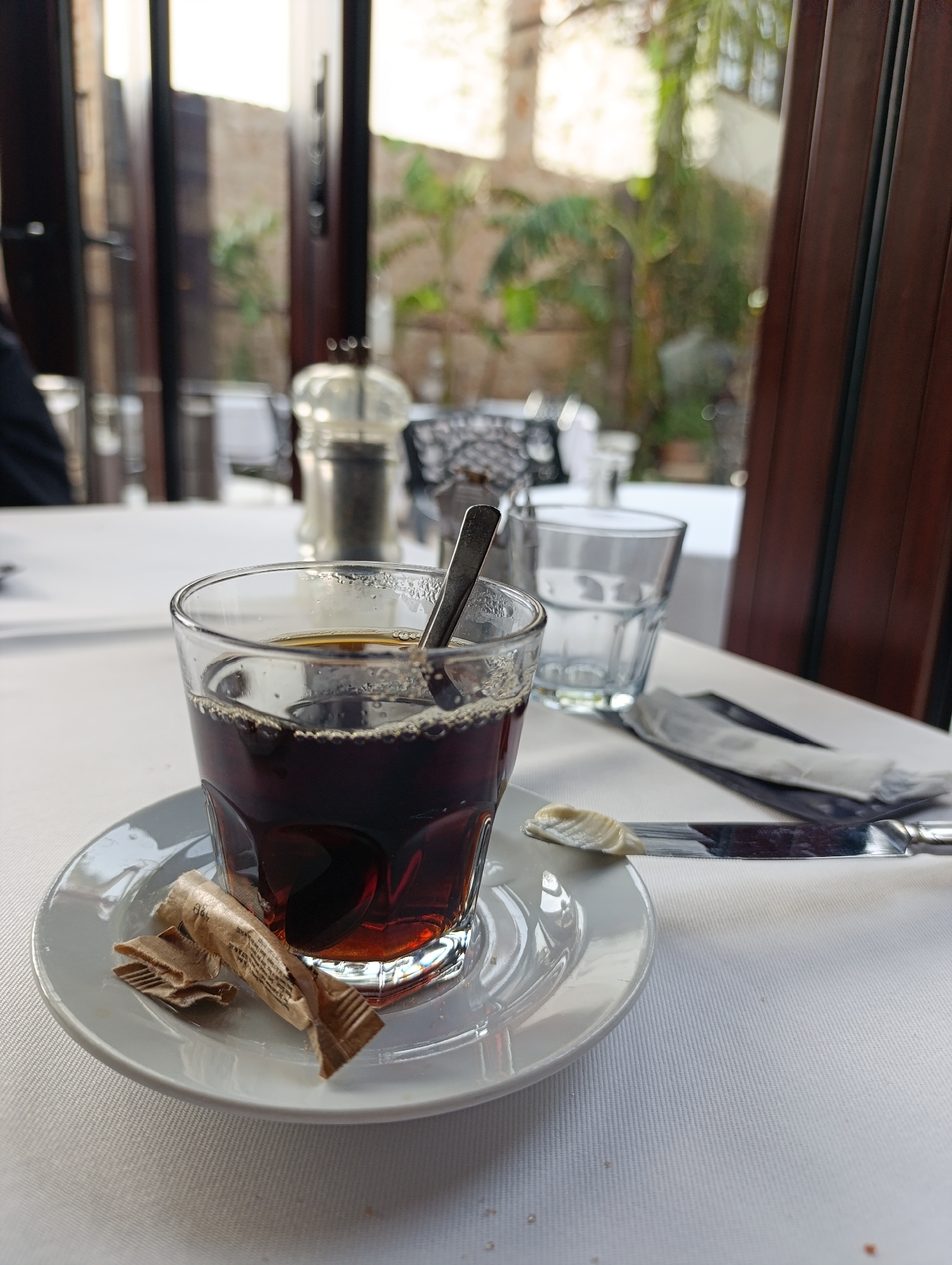 A table setting is presented indoors, possibly at a restaurant in Antalya, Turkey, given the architecture visible through the windows. The table is covered with a white tablecloth. In the foreground is a clear glass containing a dark liquid, likely coffee, with a spoon inside. The glass sits on a white saucer alongside an opened sugar packet. To the right of the coffee is a knife with a small amount of butter on the blade, and wrapped cutlery. A clear water glass stands slightly behind the coffee. In the background, there are other tables, condiments, and blurred glimpses of outdoor foliage and buildings through large windows or doors. The overall lighting suggests daytime.