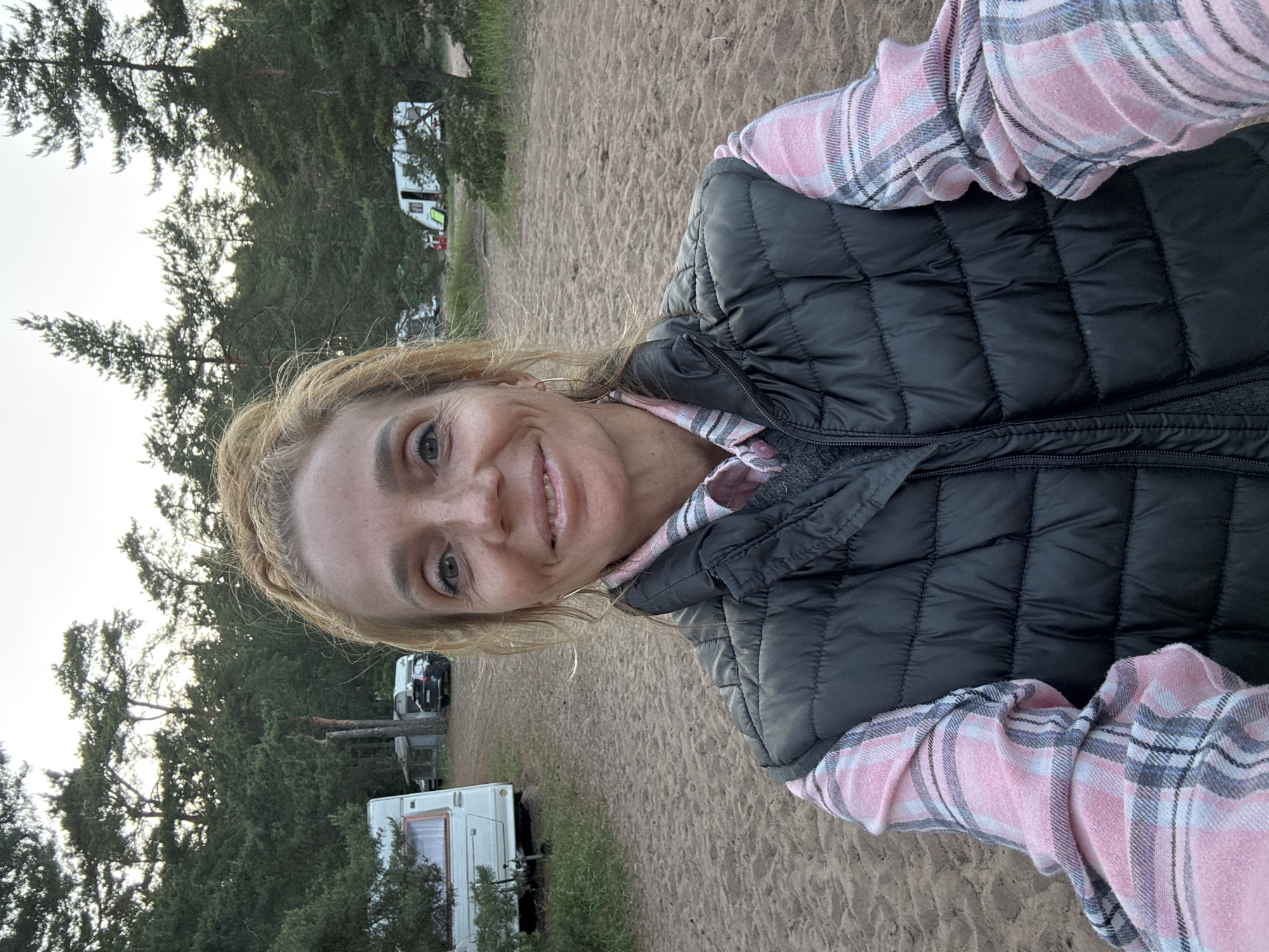 A woman with blonde hair tied up in a bun takes a selfie outdoors. She is wearing a pink and white plaid shirt and a black puffer vest. Her expression is neutral, with a slight smile. Behind her, a sandy campsite is visible, with several caravans and trees in the background. The location is identified as Zelenaya Roshcha, Russia.