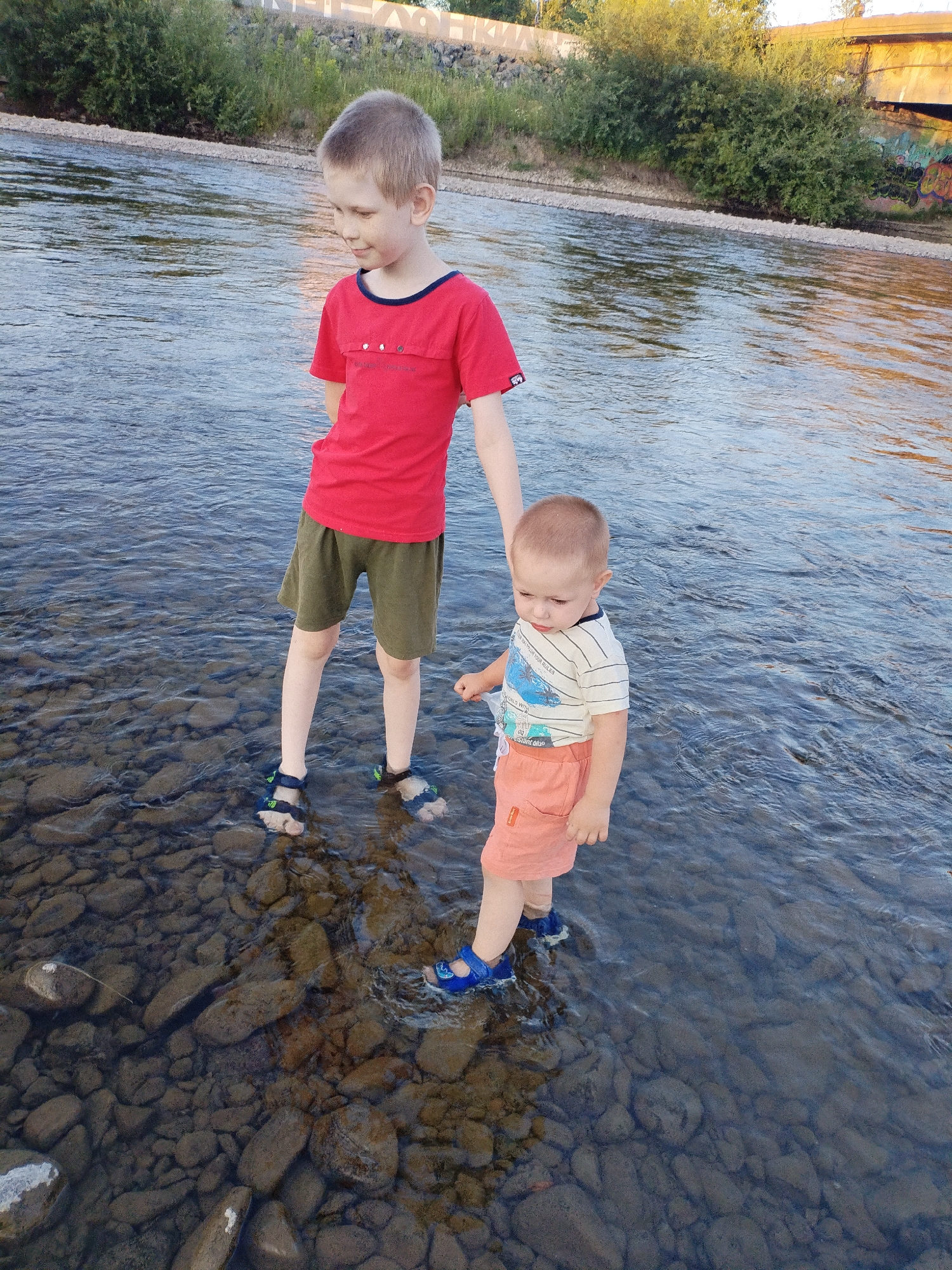 Two young boys are wading in the shallow, rocky waters of the Amur River. The older boy, on the left, is wearing a red t-shirt and olive green shorts, with dark sandals. He is looking down. The younger boy, on the right, is wearing a striped t-shirt with a blue and white graphic and orange shorts. He has blue water shoes on and is also looking down. In the background, there is a grassy bank with some trees and a concrete structure, possibly a bridge, with graffiti on it. The sun is casting a warm glow on the water. The location is Komsomolsk-on-Amur, Russia.