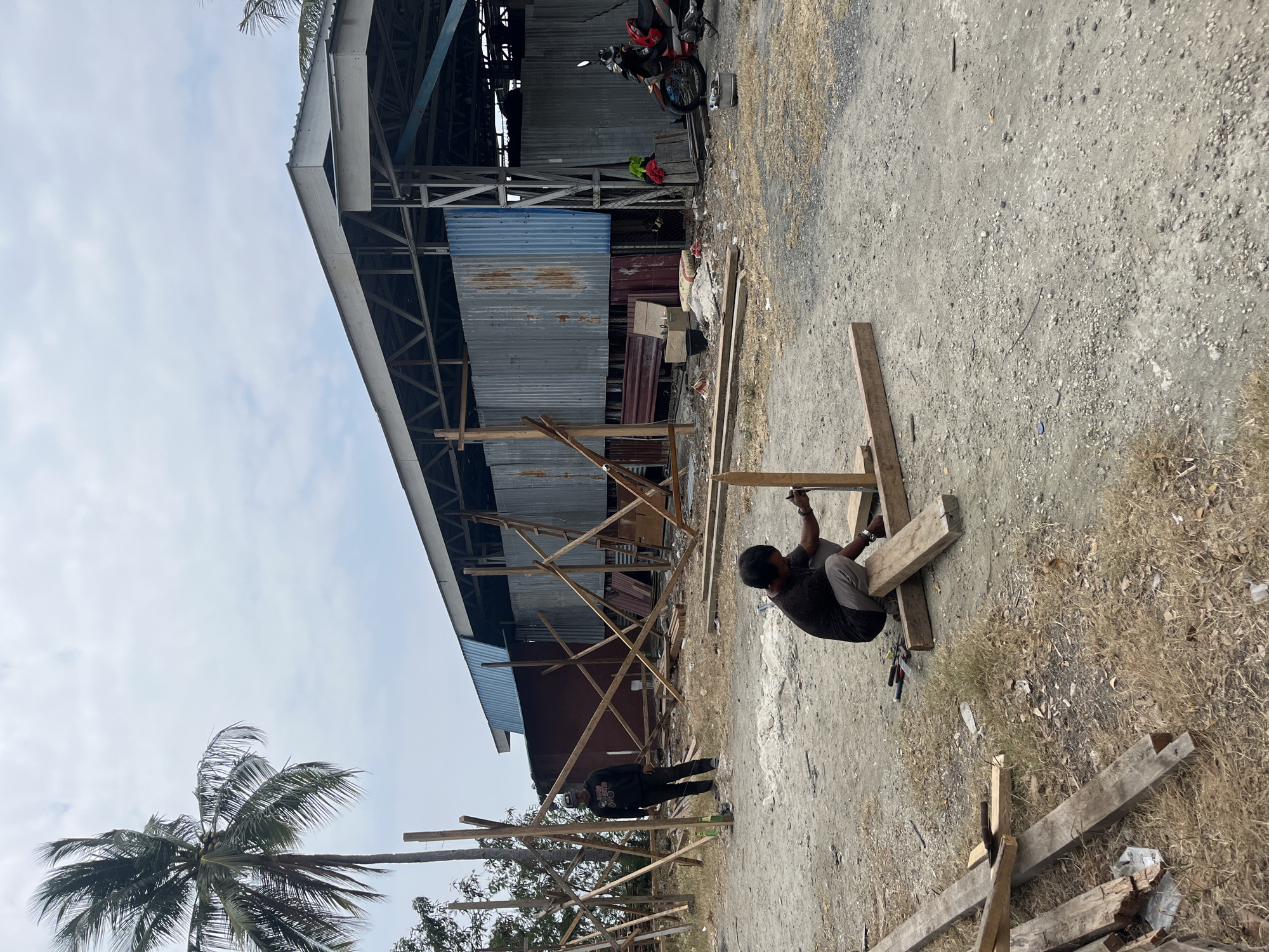Two men are working on a construction site in Luwuk, Indonesia. One man is crouching and appears to be using a measuring tape on a wooden beam. The other man is standing and watching him. The site is a dirt lot with a partially constructed building in the background. The building has a metal roof and corrugated metal walls. There are piles of wood and other construction materials scattered around the site. A palm tree is visible to the left of the frame. The sky is overcast.