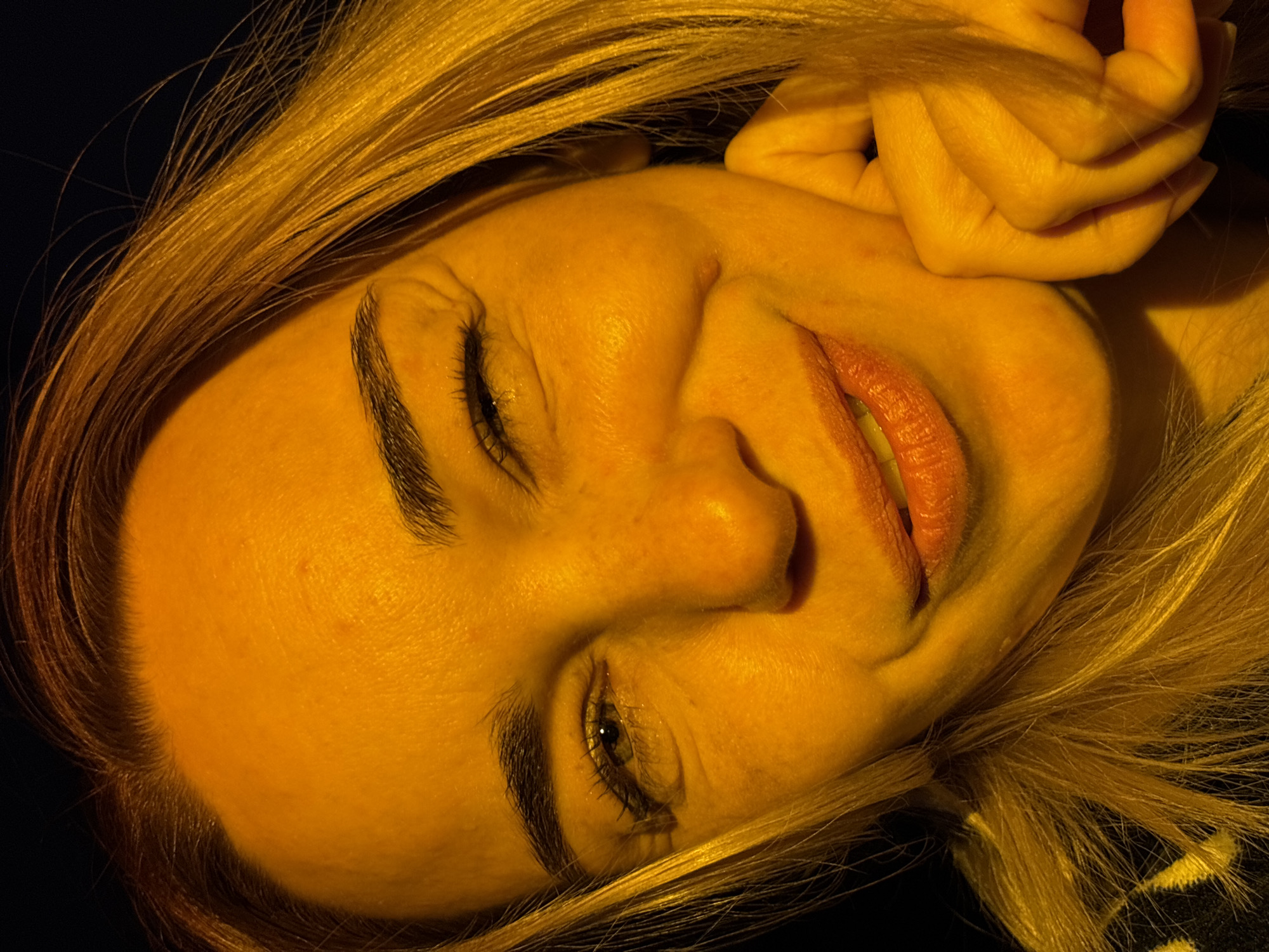 A close-up, angled portrait shows a woman with fair skin and blonde hair. Her eyes are partially closed, and she is smiling with her mouth slightly open, revealing her teeth. Her right hand is visible, holding the right side of her face near her ear. The background is dark and out of focus. The image has a warm, yellow-toned color cast. The location is Unknown, Russia.