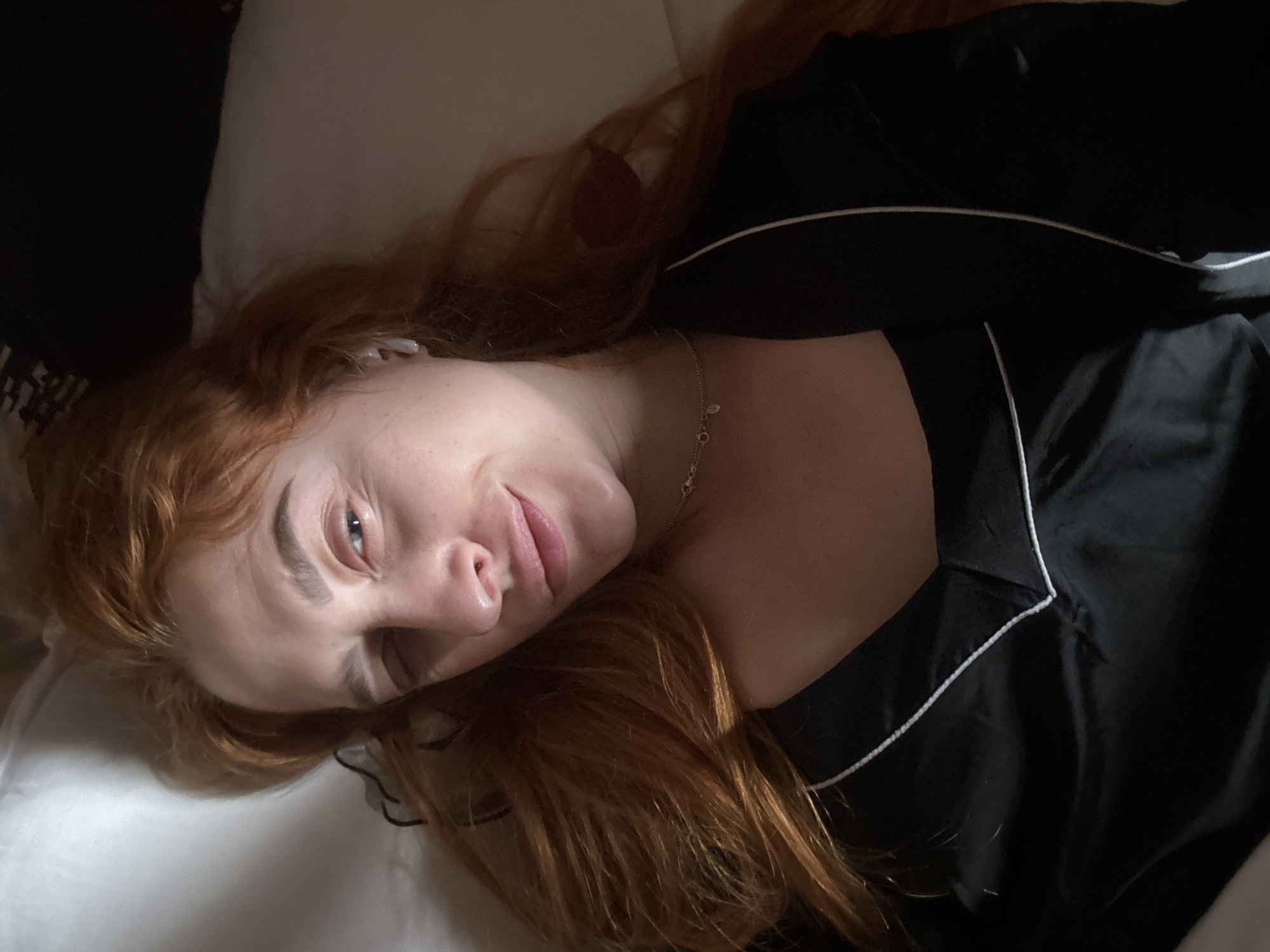 A fair-skinned woman with red hair is lying down, possibly in bed, wearing a black pajama top with white piping. She is smiling slightly and looking toward the camera. A thin, silver necklace is visible on her neck. The scene appears to be indoors. Budva, Montenegro is not discernable in the image.