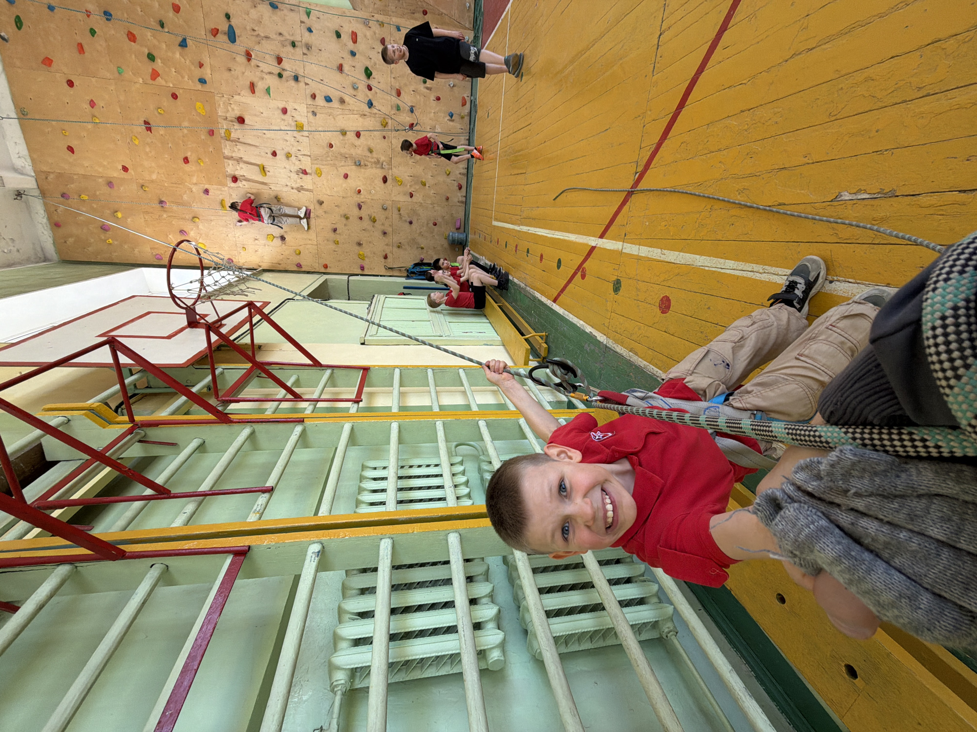 The image, taken indoors in Izhevsk, Russia, shows a group of individuals participating in rock climbing activities. In the foreground, a young boy, wearing a red shirt and harnessed, hangs upside down from a climbing rope, smiling at the camera. He is suspended near a radiator and a red metal structure, possibly part of the climbing apparatus.
In the background, against a wooden climbing wall studded with colorful holds, multiple figures are visible: one man stands on the floor, and several children are either climbing or being belayed. The gym floor is yellow with white and red lines marked on it. A climbing rope stretches from the boy in the foreground to the floor in the background, suggesting a belay system.