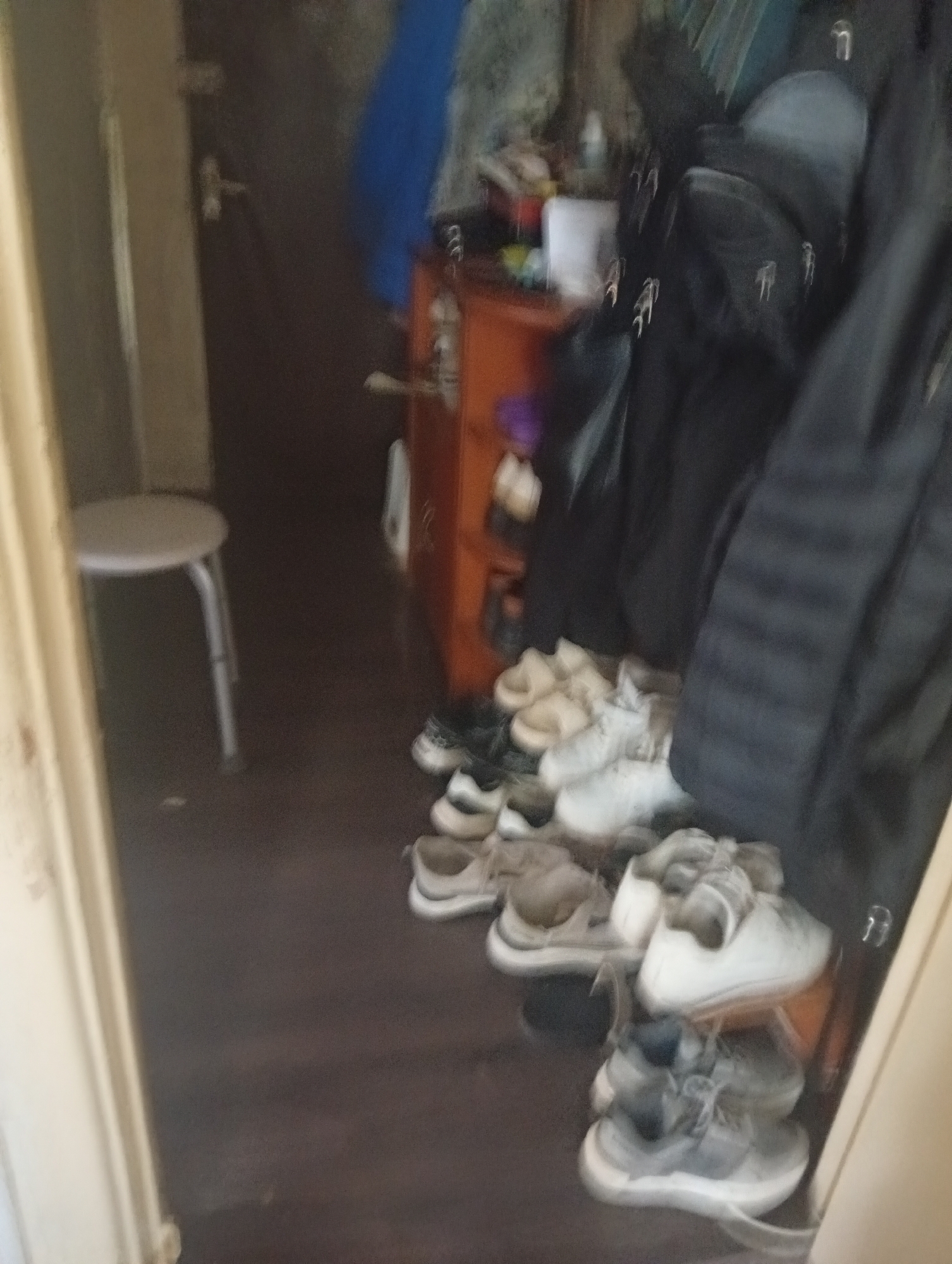 The image depicts a dimly lit hallway or entryway area, likely within a residential building in Pushkino, Russia. On the left side, a simple white stool with metal legs stands against a beige doorframe. The floor is a dark, polished wood. Towards the center and right, a collection of footwear is piled on the floor and potentially on a low shelf or rack, obscured by a dark garment. The shoes appear to be primarily sneakers and casual shoes, mostly in light colors like white and gray, with some darker pairs interspersed. A small orange cabinet or drawer unit is visible behind the shoes, featuring a silver knob. A portion of a dark blue garment or fabric is also visible further back in the scene. The overall impression is one of a functional, lived-in space with items of clothing and footwear stored in a somewhat disorganized manner. The lighting is low, creating shadows and a slightly blurry quality to the image.