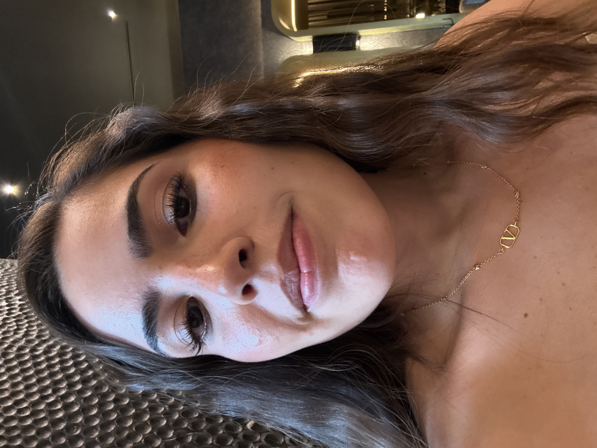 A person with brown hair and a gold necklace is lying on a textured surface. The person is facing the camera with a slight smile. The background is blurred and includes a wall with a light fixture. The location is San Bernardino Tlaxcalancingo, Mexico.