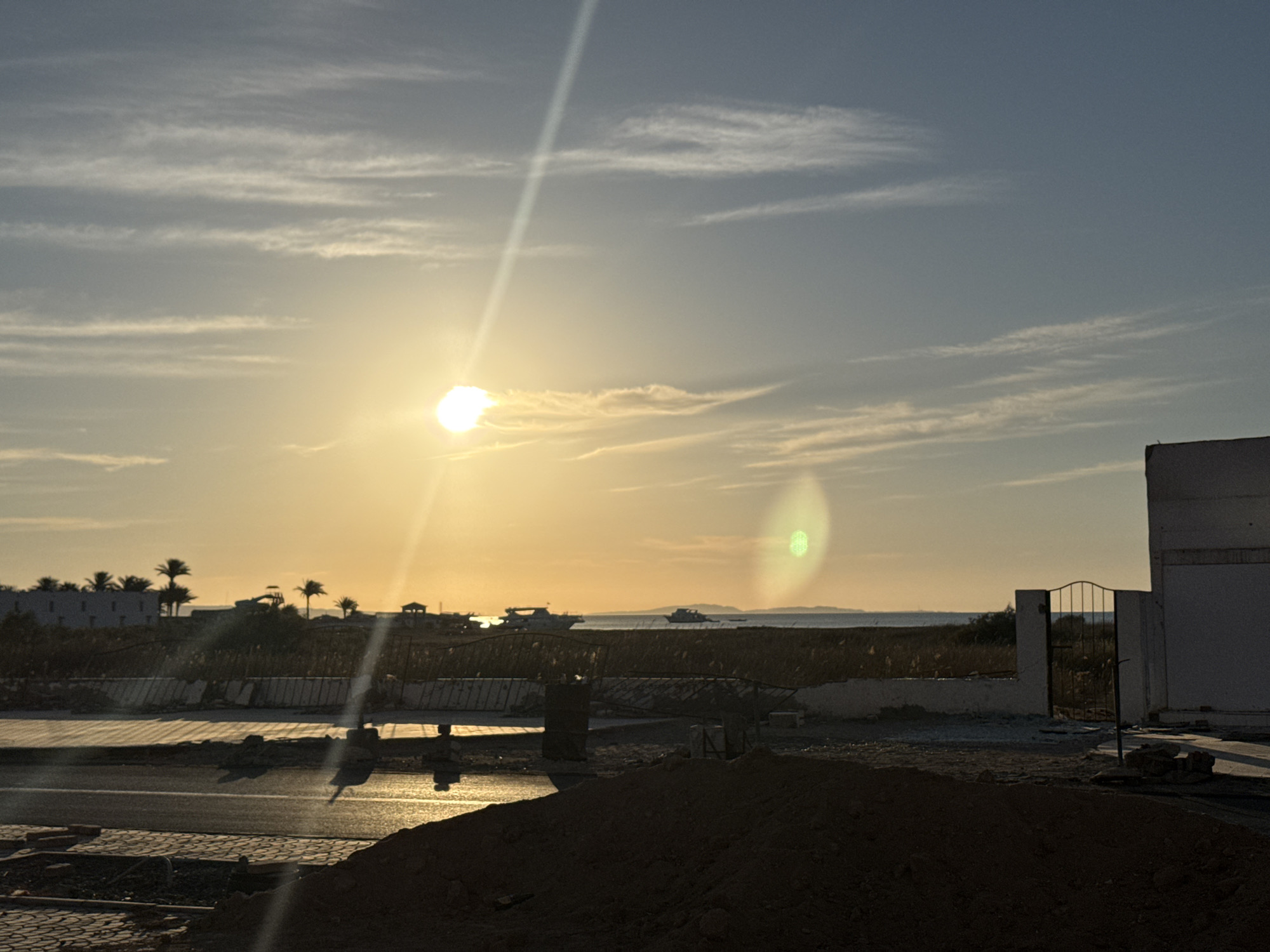 The image depicts a sunset scene, likely near a coastal area in Unknown, Unknown. The sun is low on the horizon, with a bright glare and a light streak extending upwards into the sky, which is partly cloudy. Palm trees and buildings are silhouetted in the distance. A road is visible in the foreground, with water reflecting the sunlight. There is construction debris and what appears to be a dirt pile in the foreground. A low wall with a metal gate and part of a building are on the right side of the frame. There are two boats on the ocean in the middle ground.