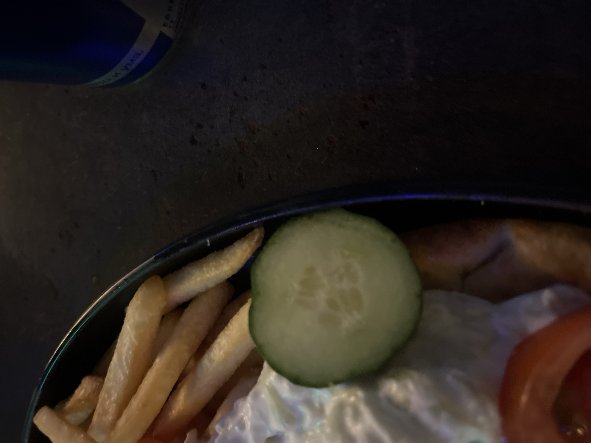 The image shows a close-up of a plate of food, including french fries, a slice of cucumber, and what appears to be a creamy white sauce or dressing, possibly with tomatoes visible below. The food is served in a dark bowl or plate. In the background, a dark, textured surface is visible, and a portion of a blue container with some text in a foreign language is also present. The lighting is dim, creating shadows and highlighting the food. There is no visible indication of the location being in the city of Plovdiv, Bulgaria; the image itself does not contain any identifiable landmarks or contextual cues that would place it in that specific city.