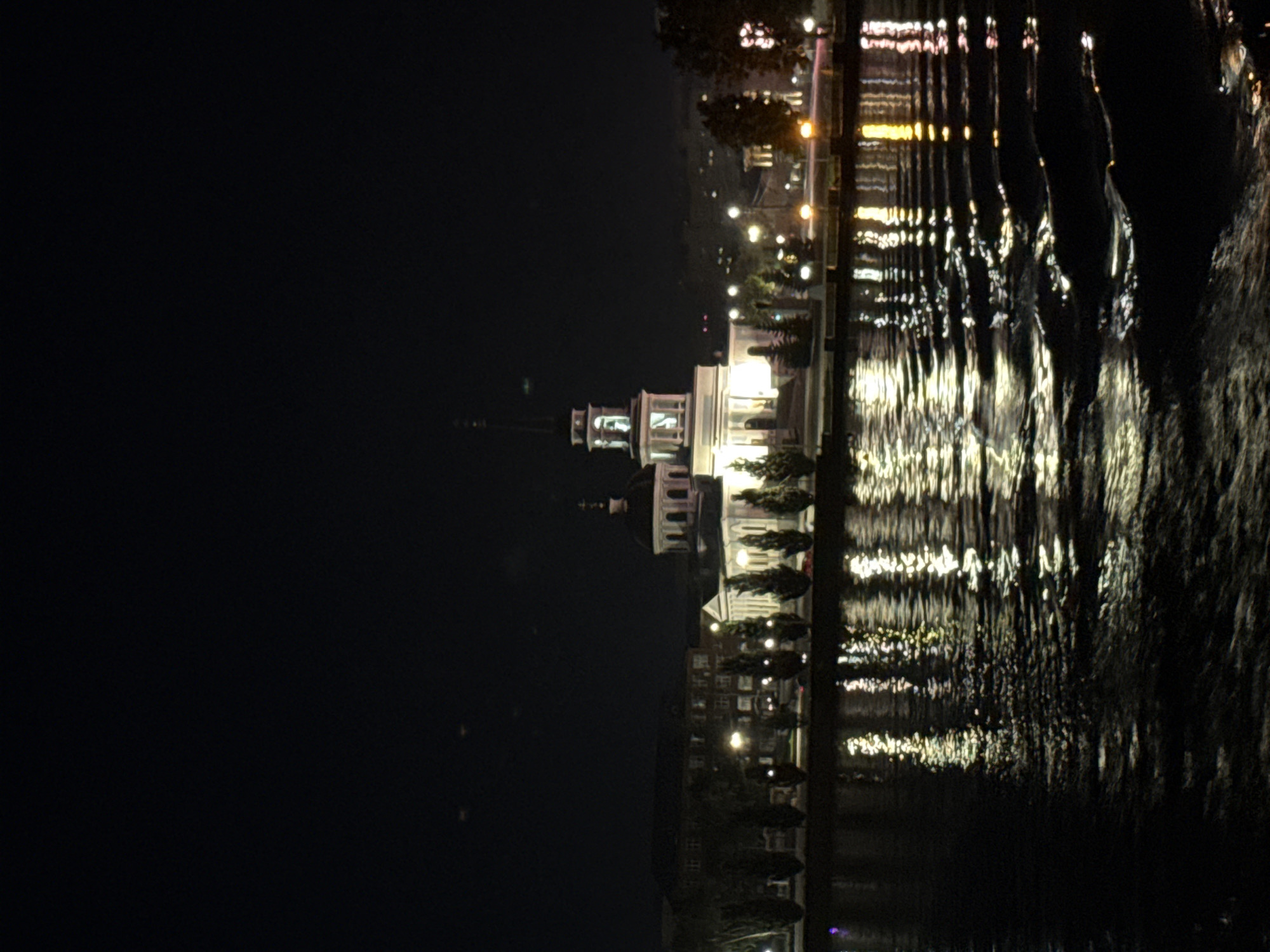 The image shows a night view of the Church of the Intercession in Votkinsk, Russia, reflected in the Votkinsk pond. The church, an architectural monument, is illuminated and its dome and spire are visible against the dark sky. The pond's surface is rippling, creating distorted reflections of the church and surrounding lights. The embankment features a row of evenly spaced trees, and beyond that, a large building complex is visible, also lit up. The scene conveys a sense of tranquility and architectural beauty at night.