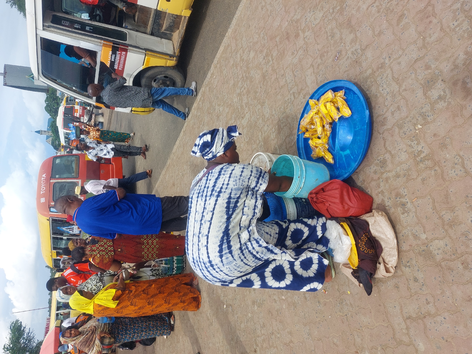 Captured from an elevated viewpoint, the image presents a bustling scene typical of a street in Dar es-Salaam, Tanzania.
The focus is on a woman dressed in a tie-dye patterned outfit, kneeling beside a blue plastic tray piled high with yellow wrapped food items, presumably for sale. Beside her are several covered containers, indicating the tools of her trade.
Around her, a group of people are gathered, seemingly waiting near red and yellow minibuses (dala-dalas), a common mode of transport in the city. The passengers are diverse in clothing, ranging from casual wear to vibrant, patterned fabrics.
In the background, more vehicles and buildings are visible, culminating in a distant view of a church steeple. The location appears to be a transport hub with a mix of pedestrian and vehicular traffic. The ground is paved with a textured surface.