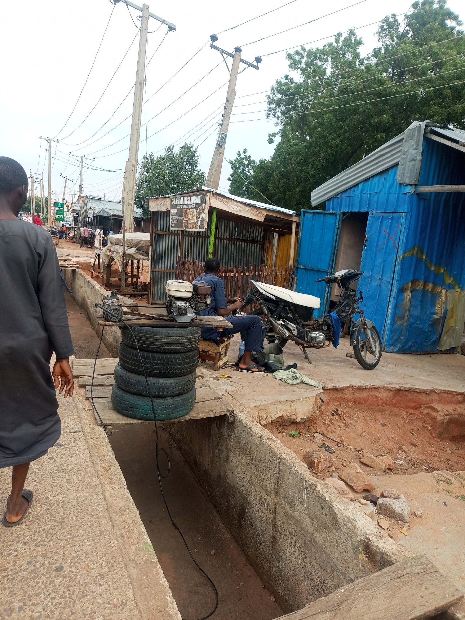In Gombe, Nigeria, on a cloudy day, a scene unfolds near a roadside business. The foreground features a concrete ditch bordering a dirt path, with a person in dark clothing walking away from the camera. Adjacent to the ditch is a wooden platform holding a stack of four tires. On top of the tires rests a small gasoline engine and a pump. To the right, a man in a blue outfit is seated on a stool next to a motorcycle. The motorcycle, a light-colored model with a white seat, is parked on the dirt ground. Behind the motorcycle is a vibrant blue corrugated metal structure with an open doorway. Further back, other small businesses with corrugated metal roofs and wooden fencing are visible, as well as electrical poles and overhead wires. The overall environment suggests a bustling, informal commercial area.