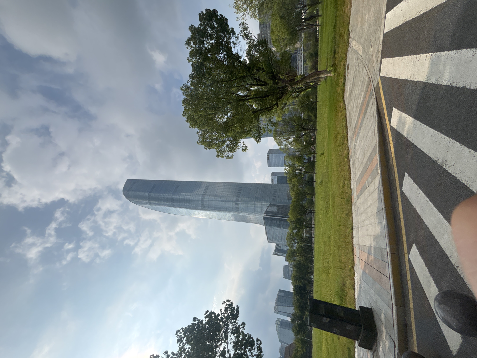 The image captures a modern cityscape with a prominent, tall, slender skyscraper featuring a curved facade dominating the center. The skyscraper is flanked by a cluster of other modern buildings with glass exteriors in the background. In the foreground, a well-maintained green lawn stretches towards the buildings, interspersed with rows of trees. A paved walkway with patterned tiles runs along the edge of the lawn, leading to a street with a pedestrian crossing. A dark, angled monument or structure is situated on the lawn near the walkway. The sky above is a mixture of blue with scattered white clouds, suggesting daytime with fair weather. This scene is located in the Suzhou Industrial Park, China, a hub for modern architecture and urban development. There are no people or discernible actions depicted in the image.