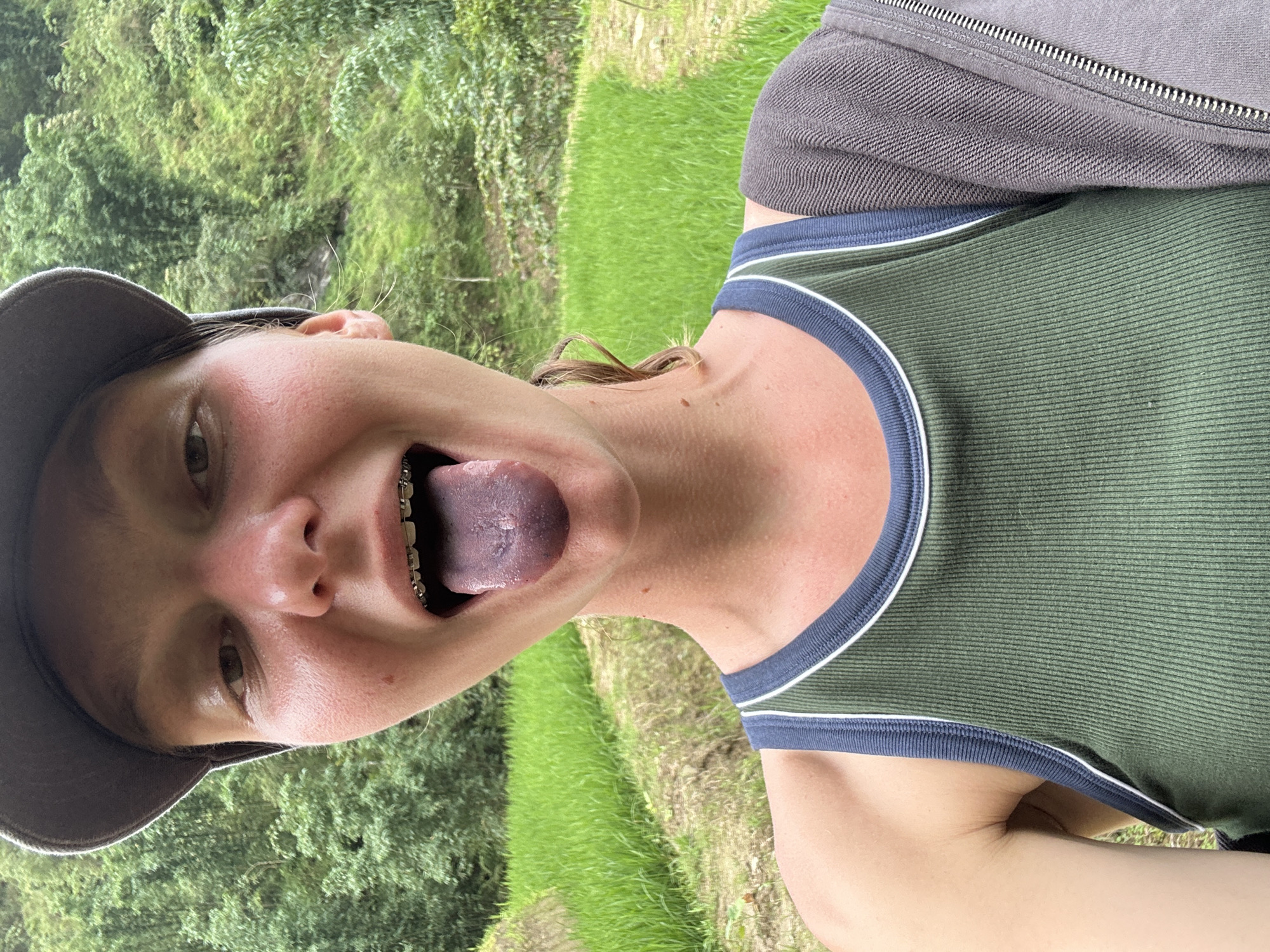 A close-up, angled shot depicts a person outdoors. The person has light skin, a cap, braces, and a dark-colored tongue. They are wearing a green tank top with blue and white trim and a gray jacket. The background features greenery, indicative of a rural area. Based on the city name provided, the location is Ban Ho, Vietnam.