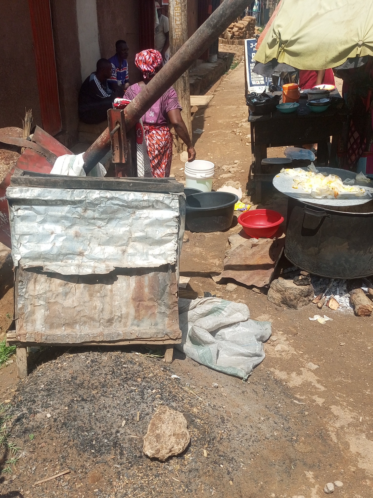 The scene is in Sabon Tasha, Nigeria, depicting an outdoor cooking and commercial activity area. A woman wearing a patterned headwrap, purple shirt, and patterned skirt is standing near a rudimentary press and carrying a white bucket. Beside her is a stand containing a pot on a stone hearth. The pot is filled with food. Beside that are plastic bowls. Nearby there is a metal bin sitting on top of a pile of ash. A white sack bag sits on the ground nearby. Further into the background, two men sit outside a building with red trim. A makeshift food stall is situated under a green-and-yellow umbrella.