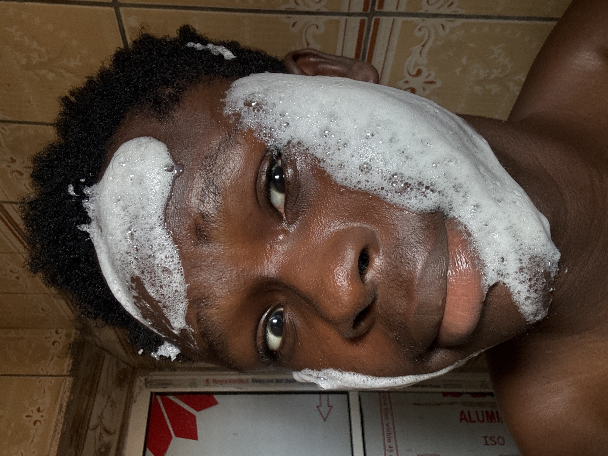 An eye-level shot captures a person, likely male, with dark skin and short, curly black hair, looking directly at the camera. The image is rotated, showing the person's head tilted to the left. White, bubbly foam covers significant portions of their face, including the forehead, cheeks, and around the nose and mouth, suggesting they are in the process of washing their face or using a facial cleanser.
The person's dark eyes are open and clearly visible through the foam, with a neutral to slightly amused expression, hinted by a subtle upturn at one side of the mouth. Their facial features, including their nose and full lips, are discernible. The upper part of a bare shoulder or chest is visible on the right side of the frame, indicating they might be shirtless or wearing dark clothing that blends with their skin.
The background consists of light brown or beige tiled walls, typical of a bathroom setting, with some decorative floral or abstract patterns on certain tiles. Below the person's chin, a reflective surface is visible, possibly part of a window frame or cabinet. On this surface, text is clearly legible, including "ALUMI" and "ISO," along with partial phrases such as "Always your best choice" and "Remember, Always your best choice." The overall scene suggests a personal hygiene moment taking place indoors in Ihiagwa, Nigeria.