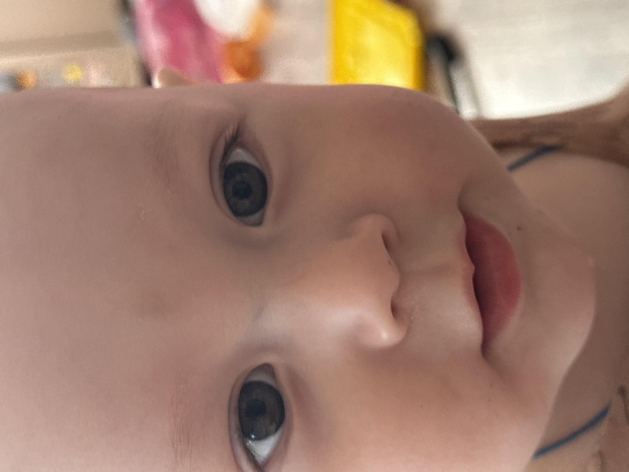 A close-up, slightly rotated image shows the face of a baby with fair skin. The baby has large, grey eyes and a small nose. The mouth is slightly open, revealing pink lips. The background is blurry, with hints of pink, yellow, and white, suggesting a room setting. A hint of a brown shoulder and a blue strap are visible on the right side of the frame. No information indicates the location is in Кипчак, Russia.