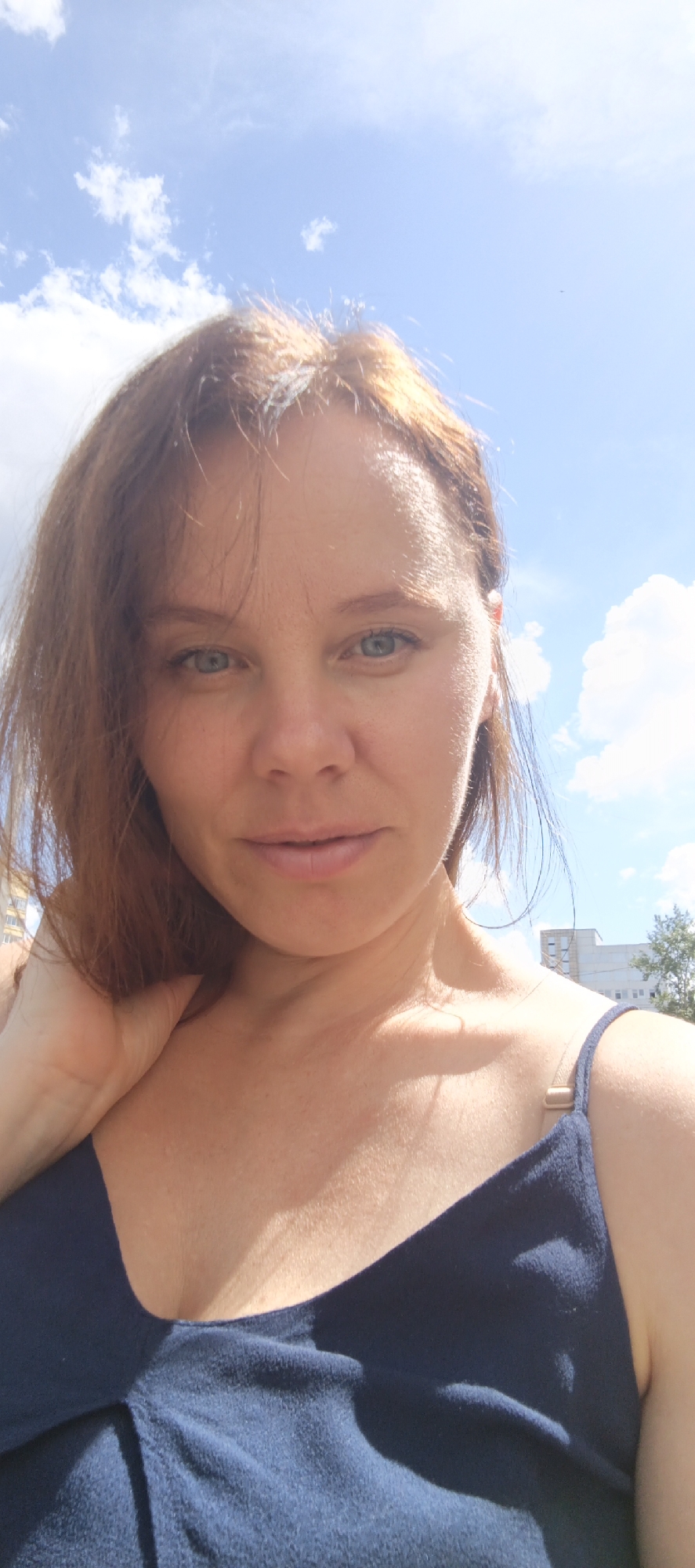 The media file is a selfie of a woman with reddish-brown hair and blue eyes, taken outdoors on a sunny day with a blue sky and scattered white clouds. She is wearing a dark blue strap top. Her left hand is touching her neck and the side of her head. In the background, there are some buildings and a tree, suggesting an urban or suburban setting. The lighting indicates it is daytime, likely mid-morning to afternoon. The woman's expression is neutral. No visible text is present. The context indicates the location is Chaikovsky, Russia.