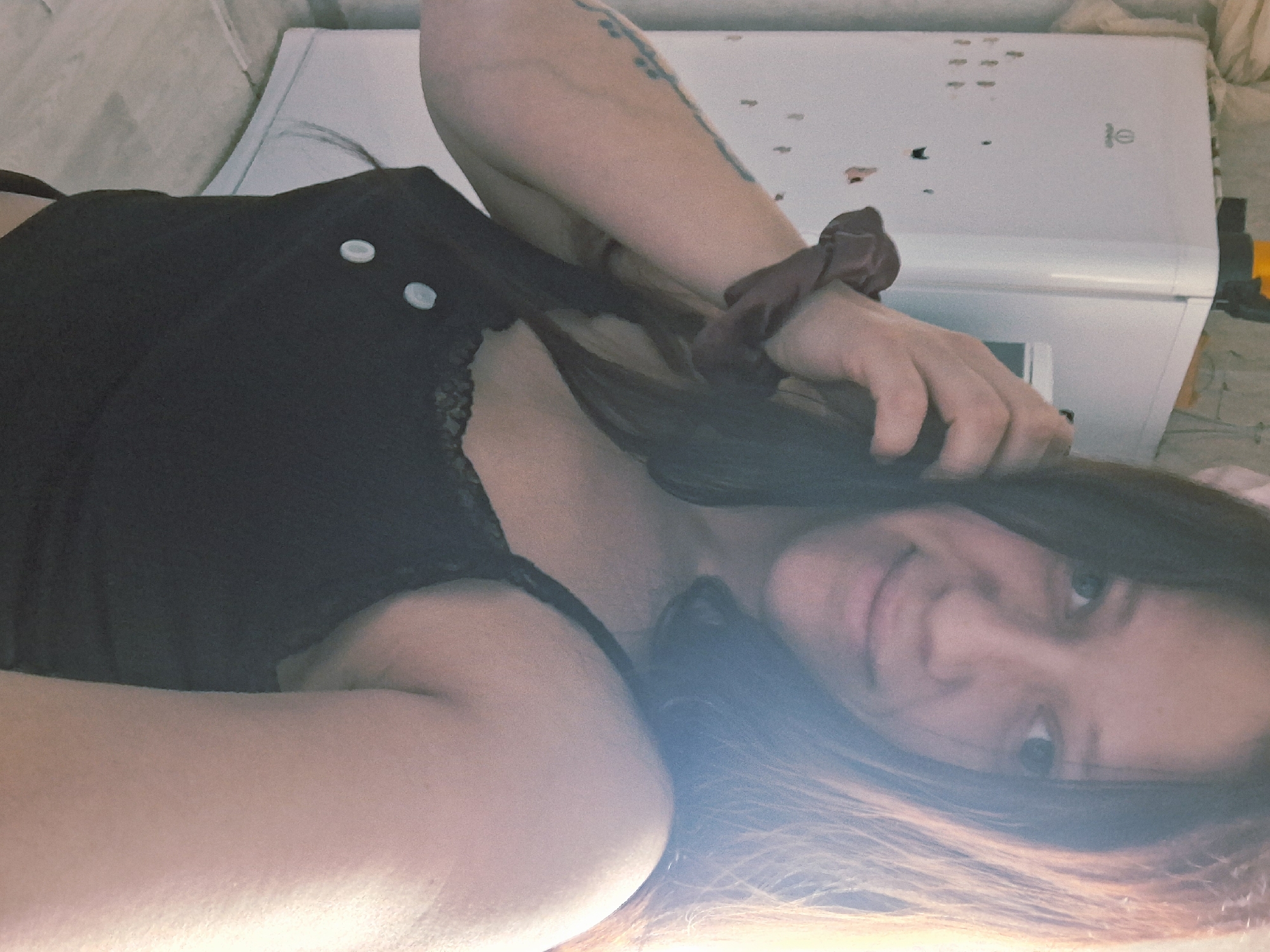 A fair-skinned person with long brown hair is lying down in what appears to be a bed. They are wearing a black top with lace trim and two white buttons. A burgundy scrunchie is on their wrist. Their arm is raised, holding some of their hair. A white appliance, possibly an air conditioner, is visible in the background. The location of Писарёвка, Russia, cannot be confirmed based on the visual information provided.