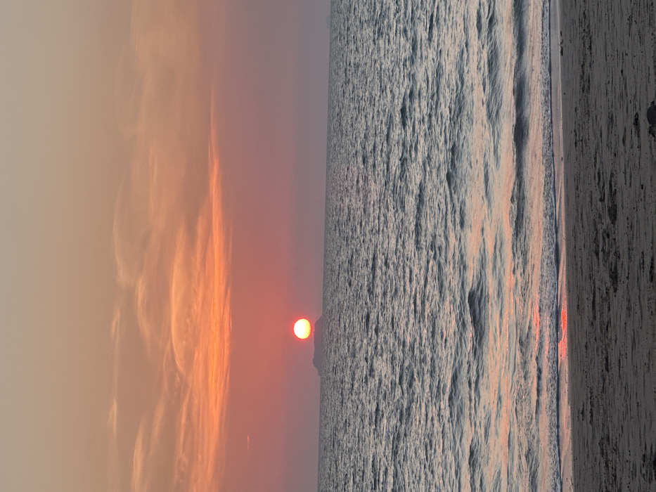 This image depicts a sunset over the ocean in Phra Ae, Thailand. The sky is dominated by hues of orange and red, transitioning from a pale peach at the top to vibrant orange and deeper red tones near the horizon. Sparse, elongated clouds are visible in the upper and middle sections of the sky. The sun, a distinct, circular, bright orange-red orb, is positioned centrally on the horizon, appearing to rest directly above a dark, low-lying island or landmass. The sea, occupying the middle ground, exhibits a dark blue-gray color with moderate ripples and reflections of the sunset's orange-pink light. In the foreground, a sandy beach, appearing dark gray, stretches across the bottom of the frame, with gentle waves breaking at its edge, creating a narrow strip of orange-red reflection where the water meets the sand. No individuals or man-made objects are present in the scene.