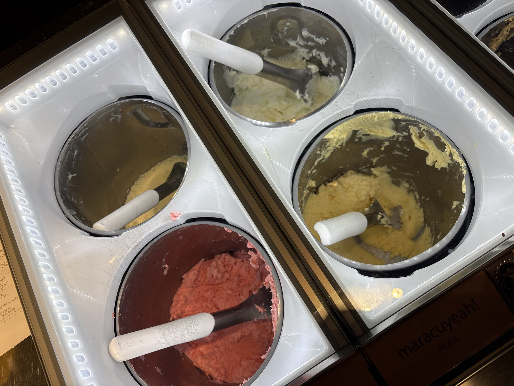 The image shows a close-up, overhead view of four stainless steel containers filled with frozen desserts, likely gelato or ice cream, displayed in a refrigerated case. Each container has a stainless steel scoop with a white handle resting inside. The desserts appear to be strawberry or raspberry on the top left, a lighter yellow, possibly vanilla or lemon, on the top right, and a more golden yellow, possibly passionfruit or mango, on the bottom right. The bottom left container's contents are partially obscured, but appear to be a yellow dessert. The display case is illuminated by a strip of white LED lights along the top edge. In the bottom left corner, a dark brown box with white text is visible, which reads "maracuyeah! PIURA". The background is dark and out of focus. Based on the text "PIURA" and "maracuyeah!", this scene is likely at a "Maracuyeah!" establishment in Piura, a city in northern Peru.