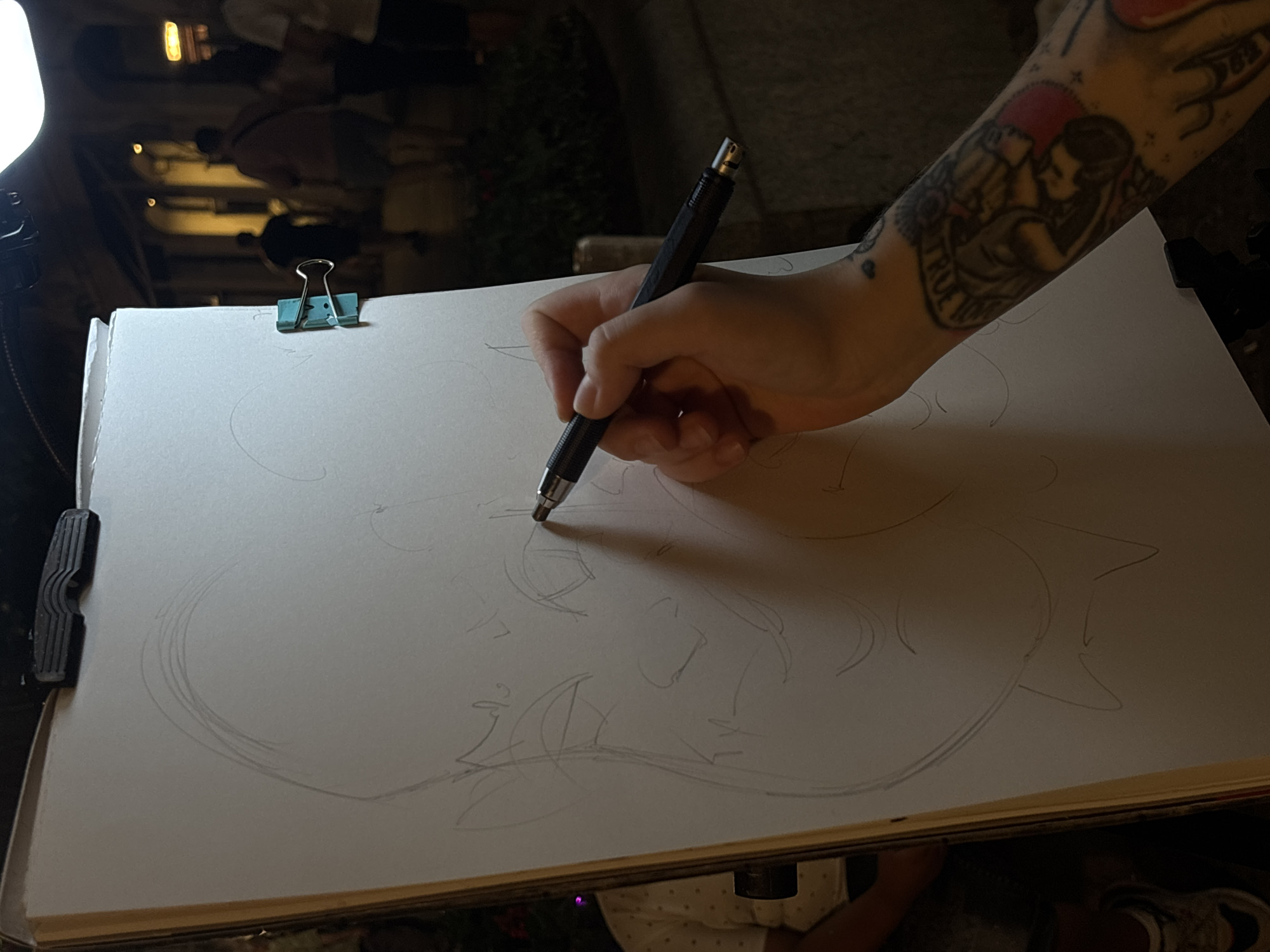 The image captures an artist's hand, adorned with tattoos, sketching on a large sheet of paper on an easel. The drawing appears to be a caricature of a face with exaggerated features. The easel is positioned outdoors, likely in a public space, as evidenced by the blurry figures of people walking in the background, some illuminated by streetlights. The setting suggests an evening or nighttime scene, given the artificial lighting and the dark, indistinct surroundings. The overall impression is one of a street artist at work, capturing a likeness in a lively urban environment. The cropped images provide closer views of the drawing details, the artist's hand and pencil, and the background setting, hinting at a bustling street or pedestrian area.
