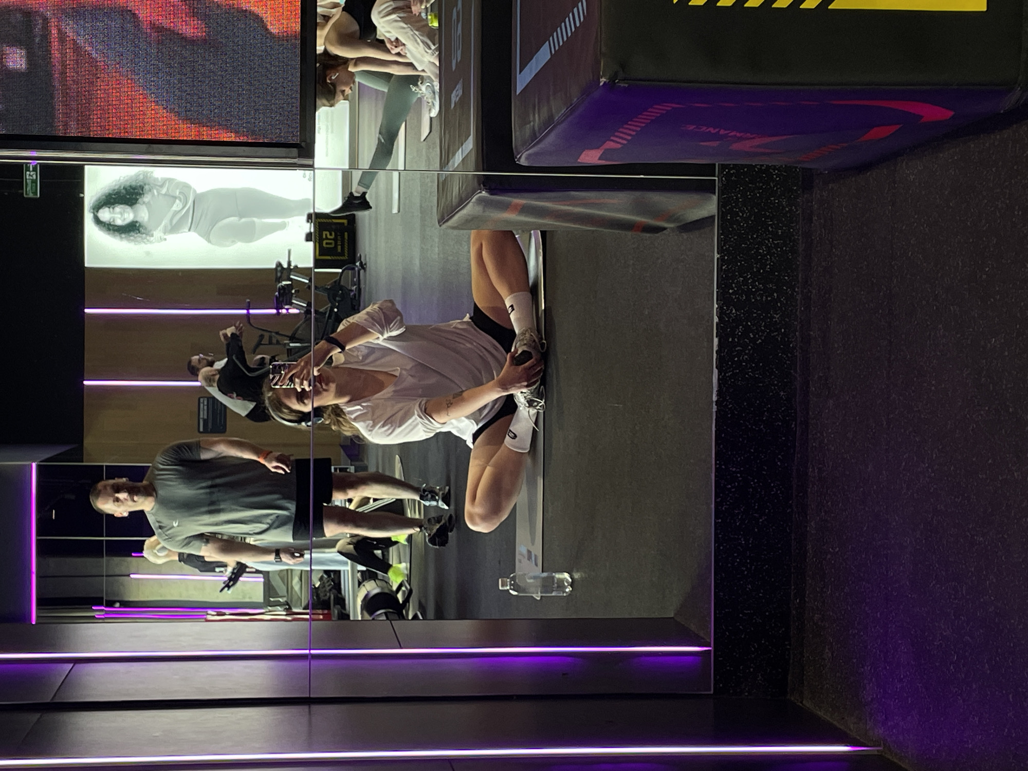 The image depicts the interior of a modern fitness studio, specifically a Performance Lab gym in Moscow, Russia, identifiable by its distinctive purple LED strip lighting, extensive mirrored surfaces, and visible branding. In the foreground reflection, a young adult female is seated on a grey yoga mat on a dark floor, performing a butterfly stretch with her legs bent and feet together. She wears a white long-sleeved shirt, black shorts, white socks, and sneakers. She has over-ear headphones on and holds a black smartphone, aiming it forward as if taking a selfie or recording. A clear plastic water bottle rests on the mat beside her. Through the mirrored background, a man in a grey t-shirt and black shorts stands centrally, facing towards the viewer. To his left, another individual is partially visible operating a stationary exercise bicycle. Further right, a person in light clothing is seen bending over. The gym features vertical purple LED light strips on the walls and an illuminated vertical panel displaying a stylized, monochromatic image of a woman in a running pose. Below this panel, a digital display shows "02". A large vertical screen on the far right displays a pixelated red and orange abstract pattern. A dark rectangular plyometric box with "PERFORMANCE" branding is partially visible in the lower right foreground.