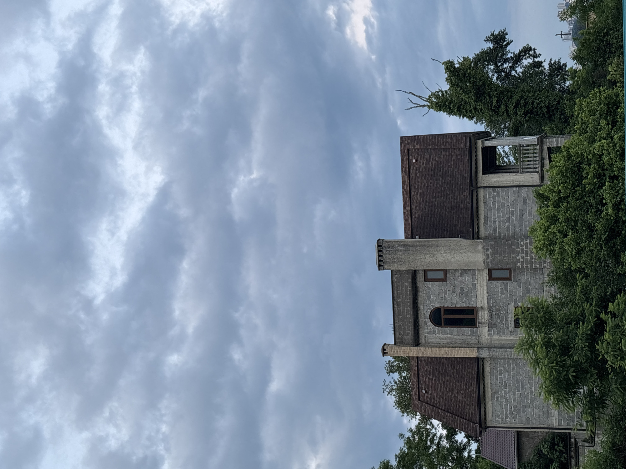 The image depicts a grey stone building with a brown roof, partially obscured by lush green foliage. The building has a large, arched window, several smaller rectangular windows, and two chimneys. There is a balcony on the right side of the structure. A tall, bare tree with twisted branches stands to the right of the building, also partially covered in green vines. In the distance, several other buildings are visible, along with a church spire. The sky above is overcast with dramatic, grey clouds. The scene appears to be a residential area in Sochi, Russia, possibly a historical or under-construction property given the unfinished appearance of the balcony. There are no people or animals visible in the image.