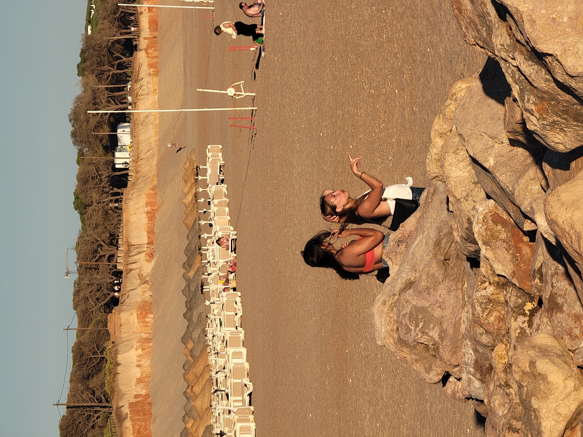 In Quarteira, Portugal, on a sandy beach with large rock formations in the foreground, two women are positioned in the middle ground. The woman on the left, with dark hair, is wearing a coral-colored bikini top and denim shorts, and appears to be adjusting her hair. The woman on the right, with blonde hair, is wearing a white top and denim shorts, and is pointing with her right hand. In the background, rows of beach chairs and umbrellas are set up on the sand. Further back, a natural cliff face with layers of sand and vegetation rises, topped by a line of trees. A few other individuals are scattered on the beach, including a woman in a white top and black pants and a man sitting on a towel. The scene is bathed in the warm light of what appears to be late afternoon or early evening, casting long shadows.