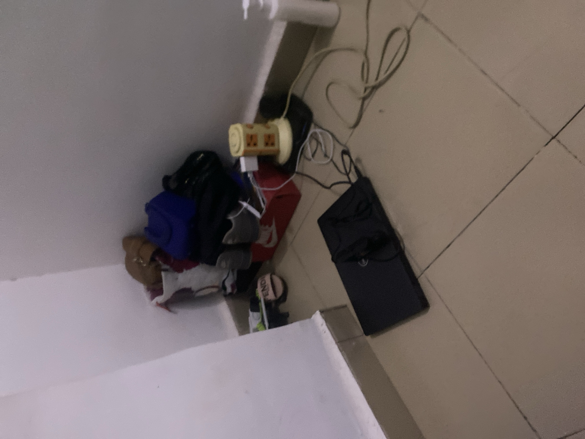 This is a photograph taken in a corner of a room in Howayi, Nigeria. The floor is tiled with light brown square tiles. In the corner, against a white wall, are several items. A stack of clothing includes a blue garment and black fabric. Next to the clothing, a red box is visible, with a pair of grey sneakers and another shoe peeking out. A tan-colored slide with the word "FENDI" is on the floor to the left of the red box. In the foreground, a black laptop is placed on the floor, connected to a charging cable that extends towards a yellow, multi-outlet power strip. A white dispenser bottle is on the floor to the right of the power strip, and various electrical cords are scattered on the floor.