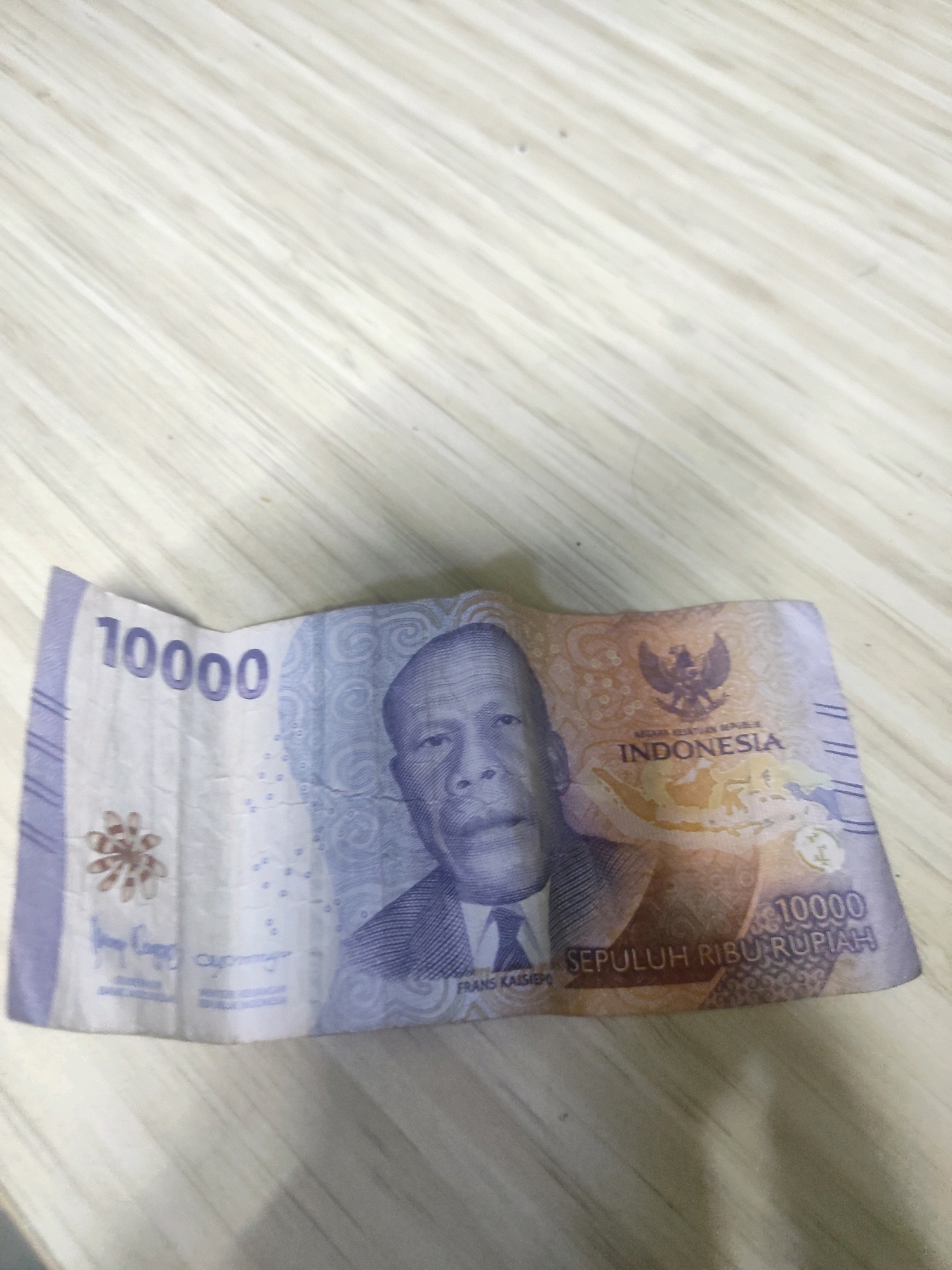 The image shows a ten thousand Indonesian Rupiah banknote, folded in the center and lying on a light-colored, wooden surface. The banknote features a portrait of Frans Kaisiepo, an Indonesian national hero. The denomination "10000" is visible on the left side of the banknote, and "SEPULUH RIBU RUPIAH" (Ten Thousand Rupiah) is written on the bottom right. The Indonesian coat of arms, the Garuda Pancasila, is also present on the banknote. The wooden surface has a natural grain pattern, with visible lines running diagonally across the image. The lighting suggests an indoor environment. There are no people present in the image, and no specific location in South Jakarta, Indonesia can be identified from the provided visual information.