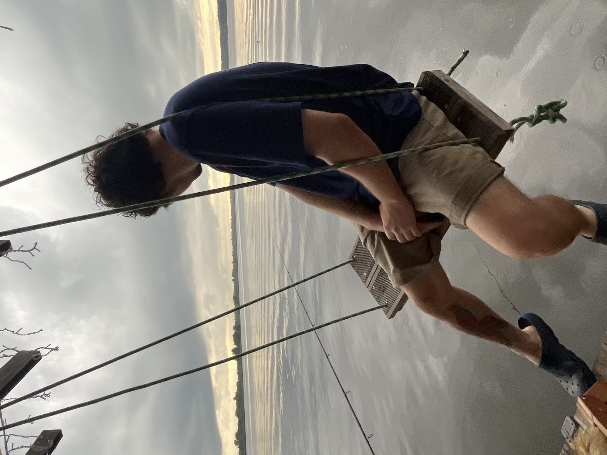 A young man with curly dark hair is seated on a wooden swing suspended over a body of water, facing away from the camera. He is wearing a dark blue t-shirt and tan shorts. His legs are extended forward, and he is wearing dark blue clogs. His left leg has a visible tattoo. Two fishing rods are positioned in the water, with their tips visible near the swing. The sky is overcast with diffused light, suggesting either dawn or dusk. The water is calm, reflecting the sky. The background shows a distant tree line. The location is identified as Radchenko, Russia.