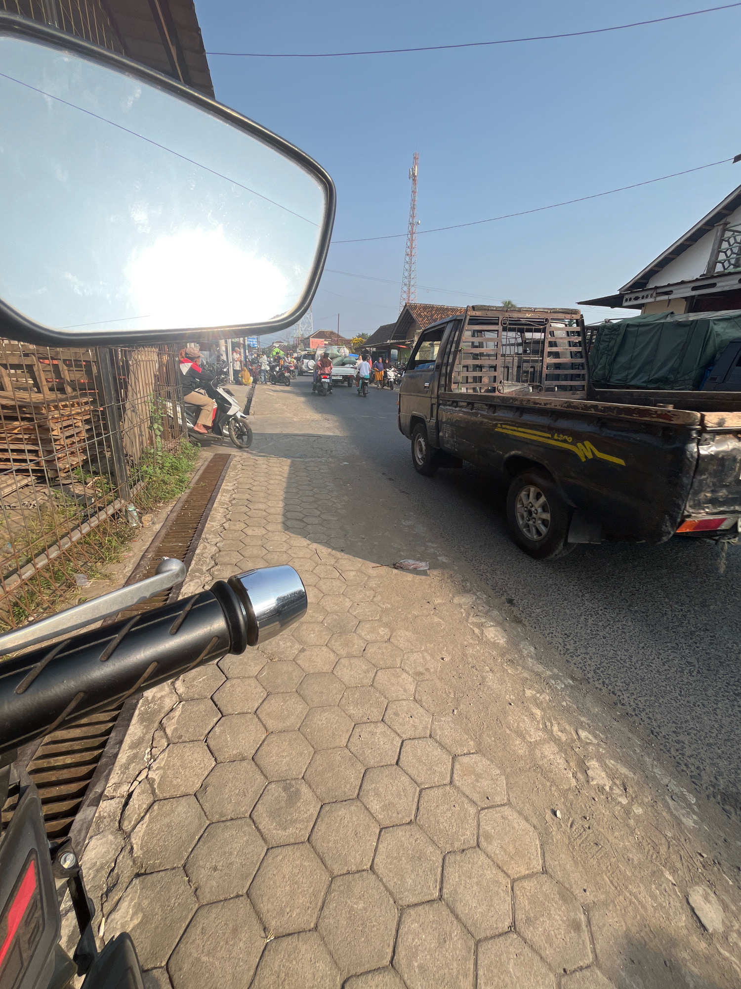 A sunny day in Kemuning, Indonesia, is depicted from the perspective of a motorcycle rider. The scene showcases a street scene with a black pickup truck with a wooden cage in the back parked on the right side of the road. The truck has yellow decals on its side. Further down the street, other vehicles and people are visible, suggesting a bustling local area. In the reflection of the motorcycle's mirror, the bright sky and some clouds are visible. The pavement in the foreground is made of hexagonal concrete blocks. A metal grate runs along the side of the road, and to the left, a fence encloses a stack of wooden planks. The overall environment appears to be a roadside in a town or village.