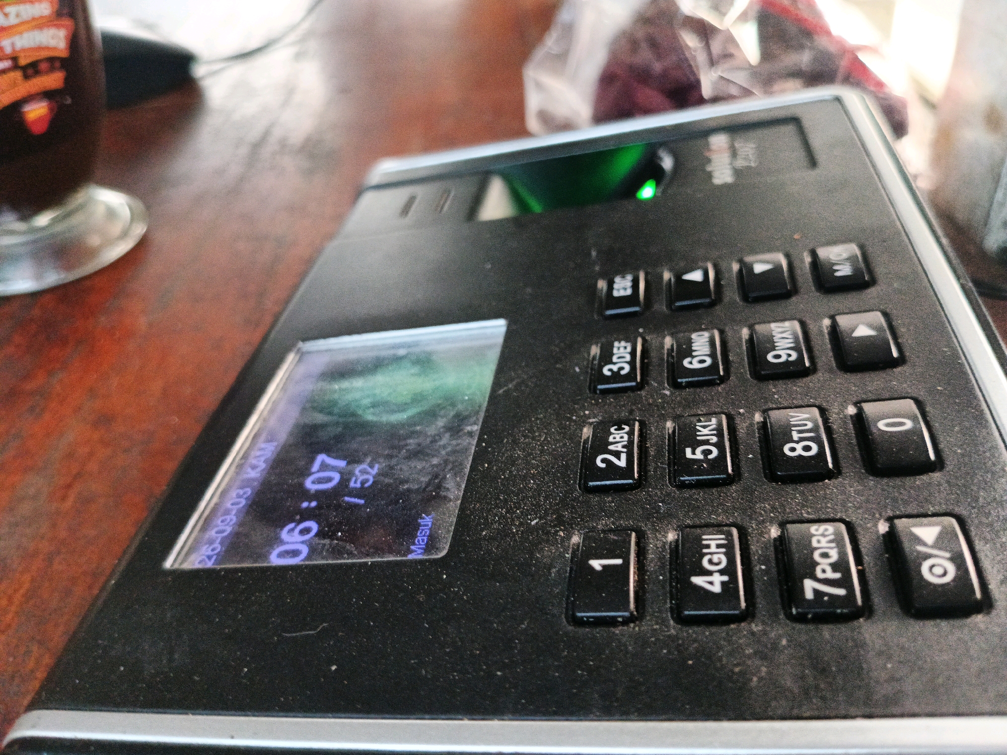 The image shows a close-up, eye-level, outdoor shot of a black fingerprint scanner device placed on a wooden surface. The device has a digital display showing the time "06:07" and the date "26-09-03". A green light is illuminated above the scanner. The surrounding area is blurred, but suggests an outdoor setting with a glass and some plastic bags containing dark objects in the background. The wooden surface appears to be a table. The image is sharp and detailed, allowing for close examination of the device's keypad and screen.