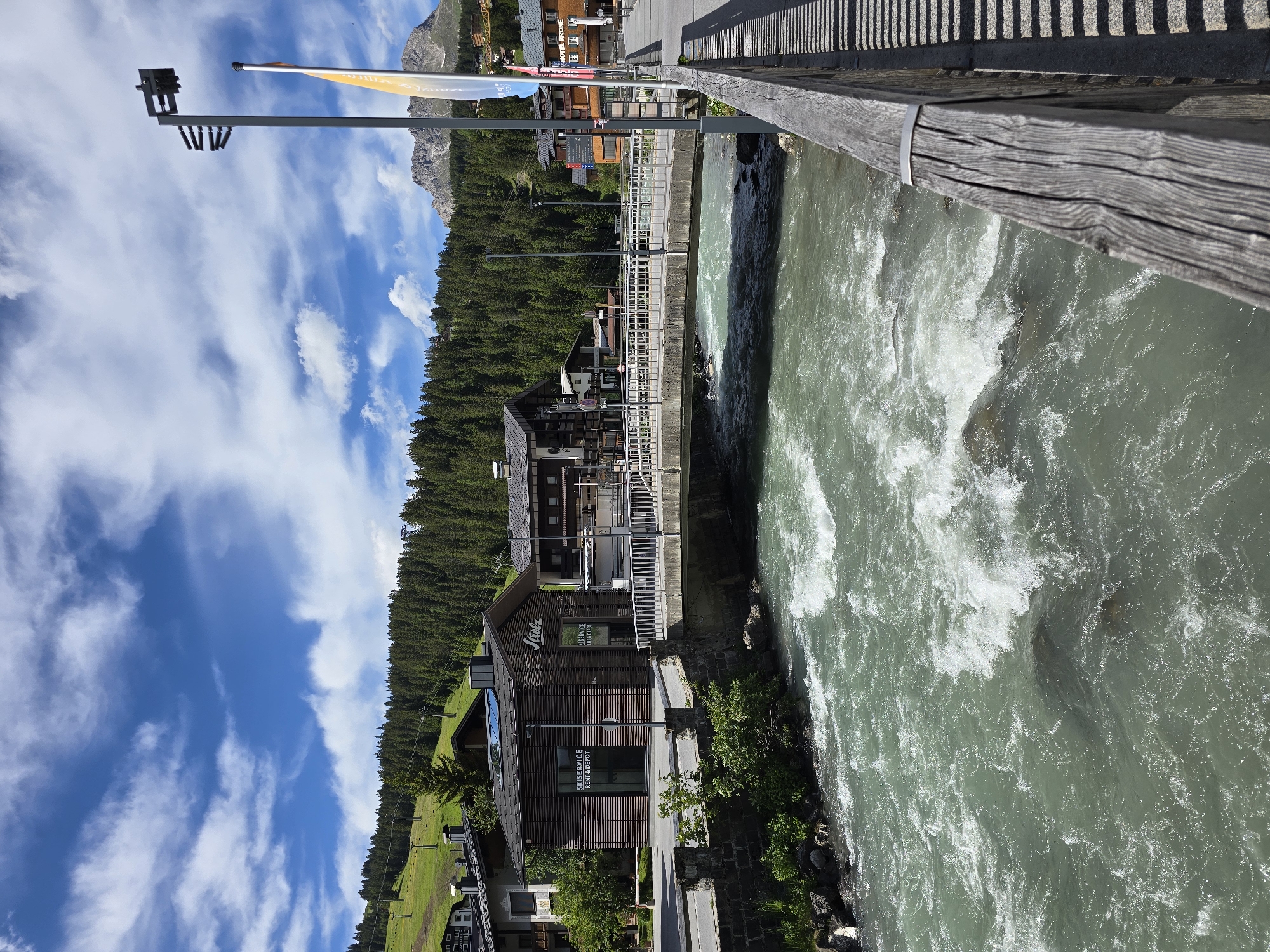 This image shows the Lech river in Lech, Austria. The river is a vibrant turquoise color and flows rapidly through the scene, with white foam indicating turbulence. A concrete bridge with metal railings spans the river in the mid-ground. On the left side of the river, a modern wooden building with large glass windows is visible, featuring the text "SKI SERVICE RENT & DEPOT" and a logo that reads "Strolz". Several other buildings, likely shops or hotels, are present on either side of the street running alongside the river. Behind the buildings, a dense forest of pine trees covers a hillside, with ski lifts visible in the upper left portion of the image, suggesting the area's connection to winter sports. The sky is blue with scattered white clouds, and in the distance, a mountain peak is visible. The foreground shows a wooden railing casting a striped shadow on a paved walkway. The overall scene depicts a picturesque mountain village during a clear, sunny day.