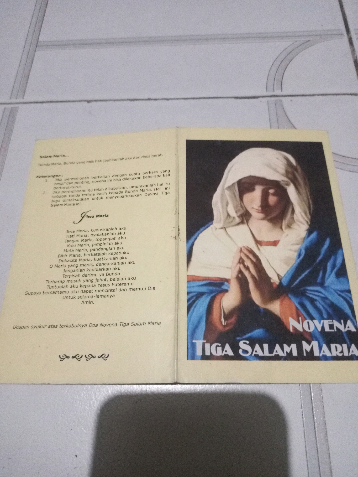The image shows an open book with religious text and an illustration of the Virgin Mary. The left page contains Indonesian text which appears to be a prayer or devotional. The right page features a large illustration of the Virgin Mary with her hands clasped in prayer, wearing a blue robe and white veil. Below the illustration, the words "NOVENA TIGA SALAM MARIA" are prominently displayed in white text. The book is placed on a white, tiled surface, likely a floor or table, with a shadow cast from the book and a portion of what appears to be a wall with vertical lines visible in the background. The overall lighting suggests an indoor setting. There is no visible indication of the city of Malang, Indonesia, or any specific geographic markers within the image.