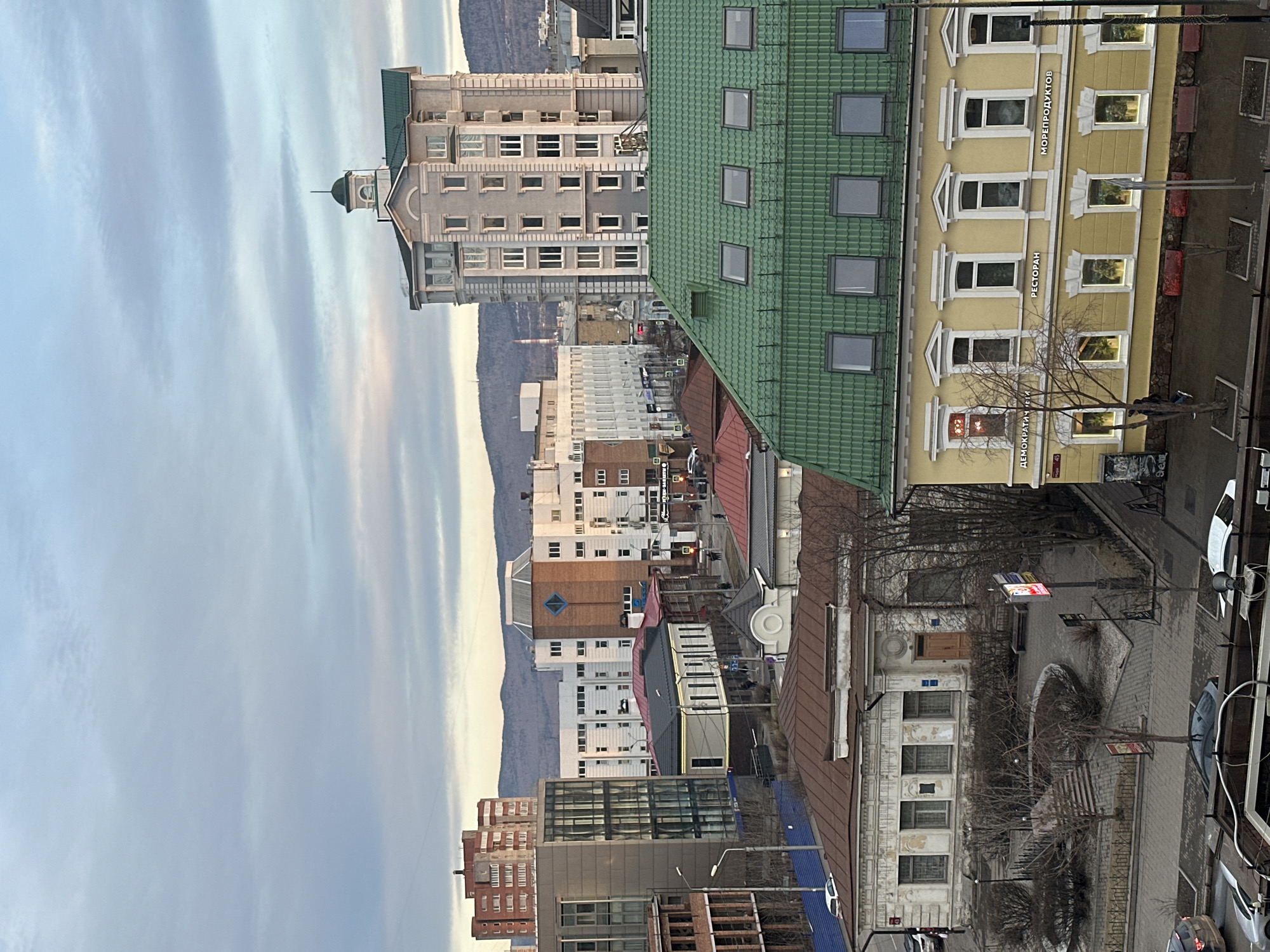 This image captures an elevated view of a city in Krasnoyarsk, Russia, showcasing a mix of architectural styles under a soft, diffused sky. The perspective looks over several buildings towards a distant mountain range.
The sky, filling the upper portion, features streaky, horizontal cloud formations, suggesting a late afternoon or early evening setting with gentle, ambient light.
In the mid-ground, a prominent yellow building with ornate white window frames and decorative trim stands out. Several windows are illuminated from within, indicating businesses. Visible text on this building reads "РЕСТОРАН" (RESTAURANT) and "МОРЕПРОДУКТОВ" (SEAFOOD), likely identifying a seafood restaurant. Further left, part of an address is visible as "ДЕМОКРАТИЧЕСКАЯ 46" (DEMOKRATICHESKAYA 46). Adjacent to it, a building with a distinctive green tiled roof and multiple skylights extends across the middle ground.
Behind these, a tall, multi-story grey building featuring a green-domed cupola or tower at its apex adds a historical or grand architectural element to the skyline. Various other residential and commercial structures, some with red or brown roofs, extend into the background, creating a dense urban tapestry. A range of hazy mountains or hills forms the distant horizon.
Below, streets are visible with parked cars, including a white vehicle on the right. Bare-branched trees, suggesting late autumn or winter, are scattered near the buildings and in a central courtyard area. No specific human activity is discernible from this vantage point.