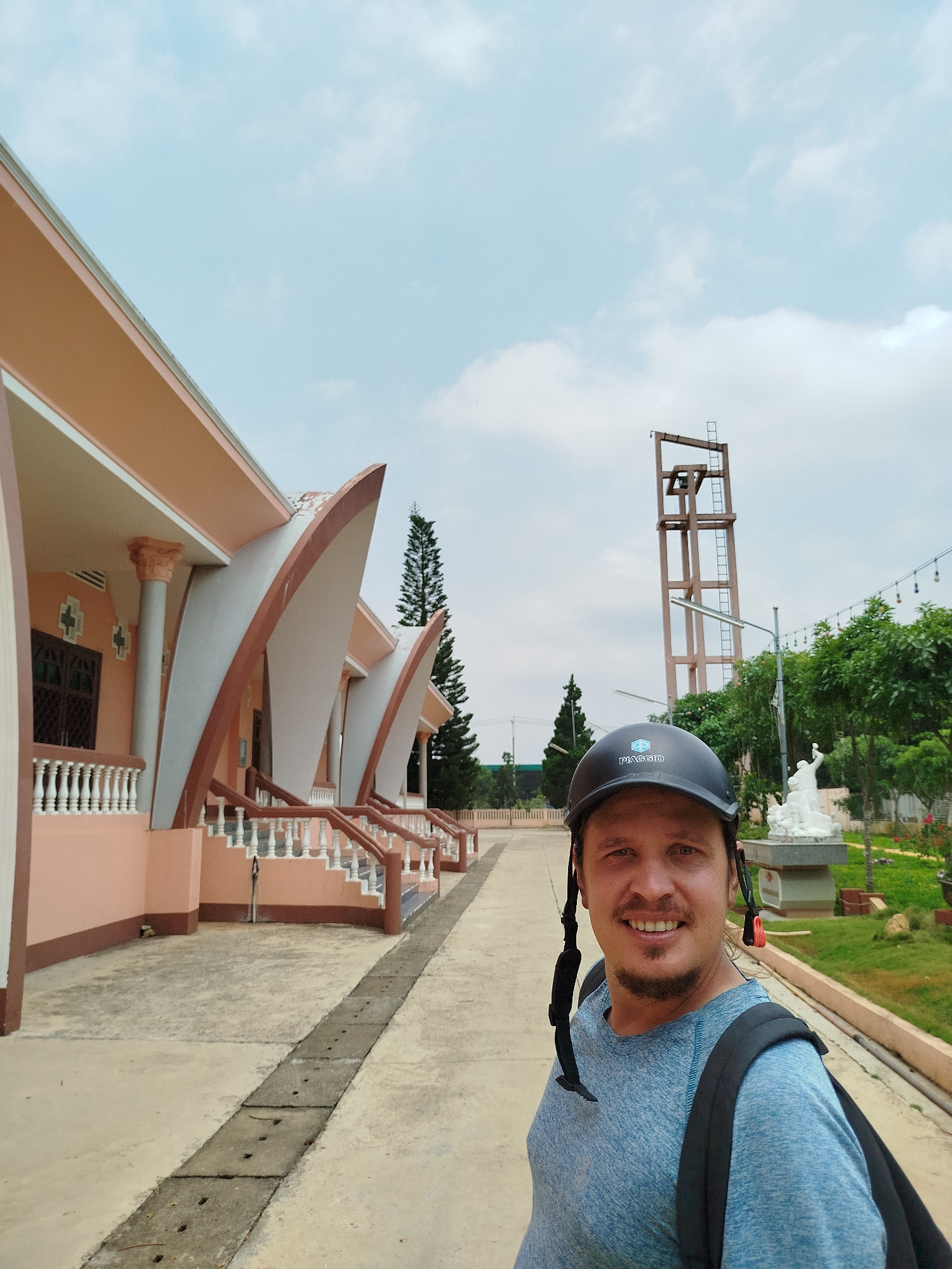 A man wearing a helmet and a backpack is standing in front of a pink building with an unusual architectural design, featuring curved, awning-like structures. The scene appears to be in Tân Hiệp, Vietnam, as the helmet bears the Piaggio logo, a common mode of transportation in Vietnam. Behind the man, there's a tall metal tower, possibly a watchtower, and a statue in a garden setting. The sky is overcast with a few clouds. The man is smiling. The area is paved and well-maintained.